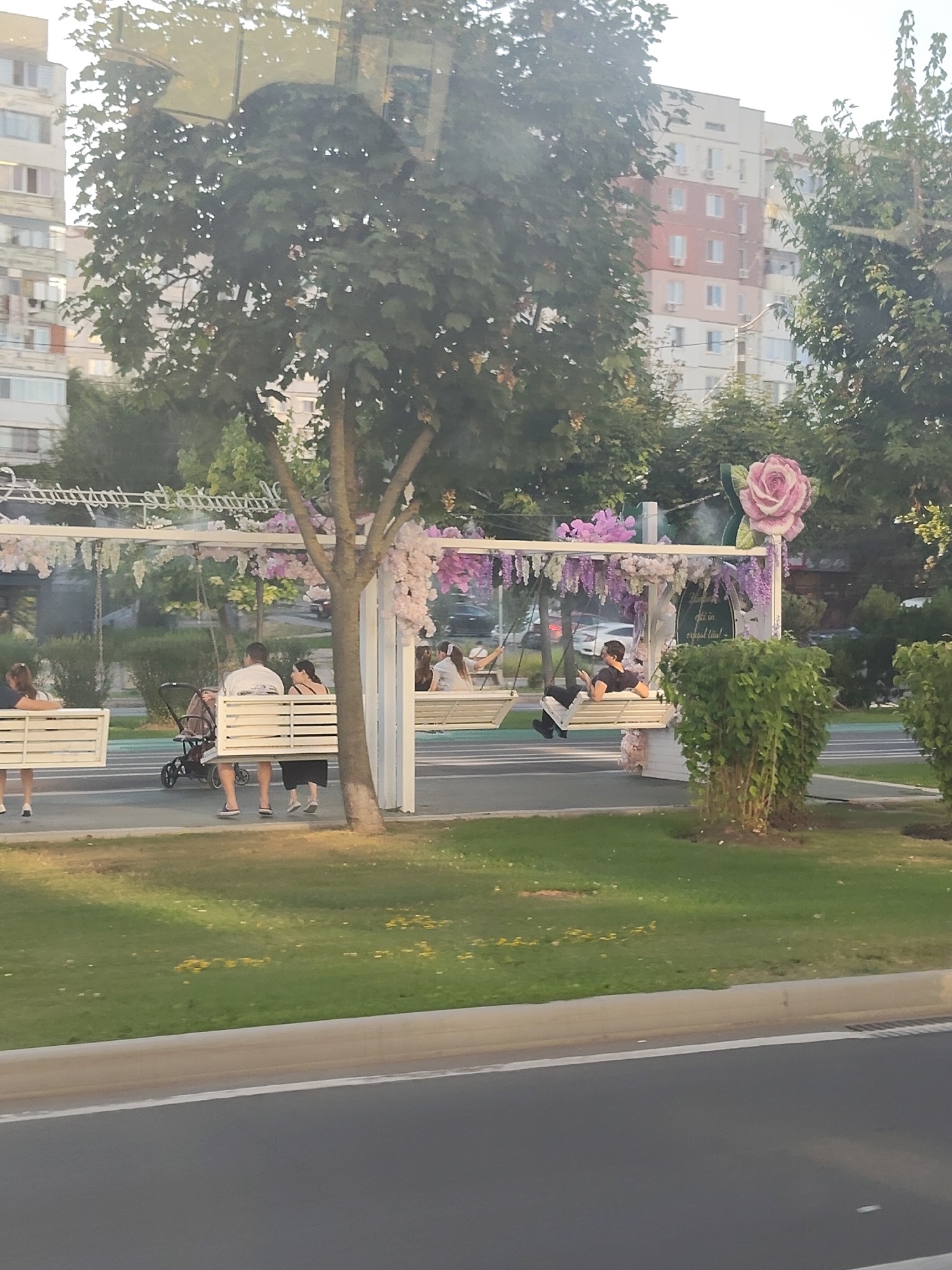 The image captures a scene in the city of Chișinău, Moldova, likely an outdoor seating area or a park. In the foreground, a section of a road is visible with a concrete curb and a grassy area bordering it. Beyond the grass, a white pergola structure, decorated with pink and purple flowers and greenery, is set up. Beneath the pergola, several people are seated on white benches or swings. On the left, a woman is seen from the back, looking to the left. Next to her, a couple is seated, with a stroller between them. Further to the right, a man is seated on a swing, looking down at his phone. Another person is visible on a swing to the far right. In the background, residential buildings are visible, suggesting an urban setting. The overall atmosphere appears to be relaxed, with people enjoying the outdoor space.