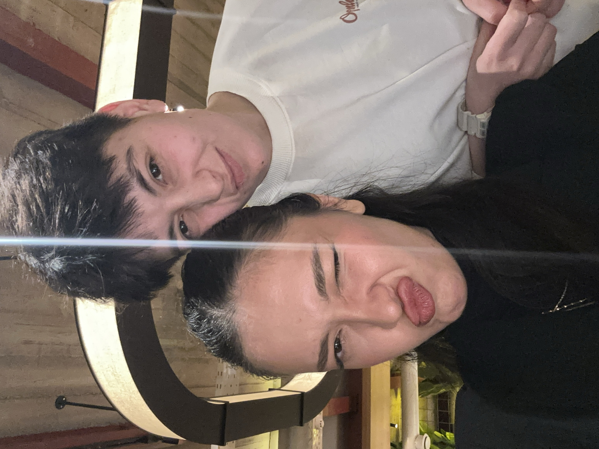 The scene depicts two individuals indoors at Lavash Restaurant in Yerevan, Armenia. A selfie perspective captures the subjects from the chest up against a warm-toned, wood-paneled ceiling with a distinctive large, curved, illuminated light fixture positioned behind them.
In the foreground, an individual with long, dark hair wears a black turtleneck-style top and a delicate silver-colored necklace with a small pendant. This individual is making a playful facial expression, puckering their lips in a "duck face" and winking their right eye.
Behind and slightly to the right, a second individual with short, dark hair wears a light-colored (off-white/cream) crew-neck sweatshirt featuring a reddish-brown script logo on the right chest, which appears to read "Orphée." This individual has a subtle smile and wears a simple, light-colored earring in their visible left ear. A light-colored, segmented-band watch is visible on their left wrist. Their left hand, with fingers interlocked, is visible near the bottom right.
A strong vertical light flare extends from the top of the frame to the lower center. The background also shows glimpses of ambient lighting and some green foliage on the far left, suggesting an upscale casual dining environment.