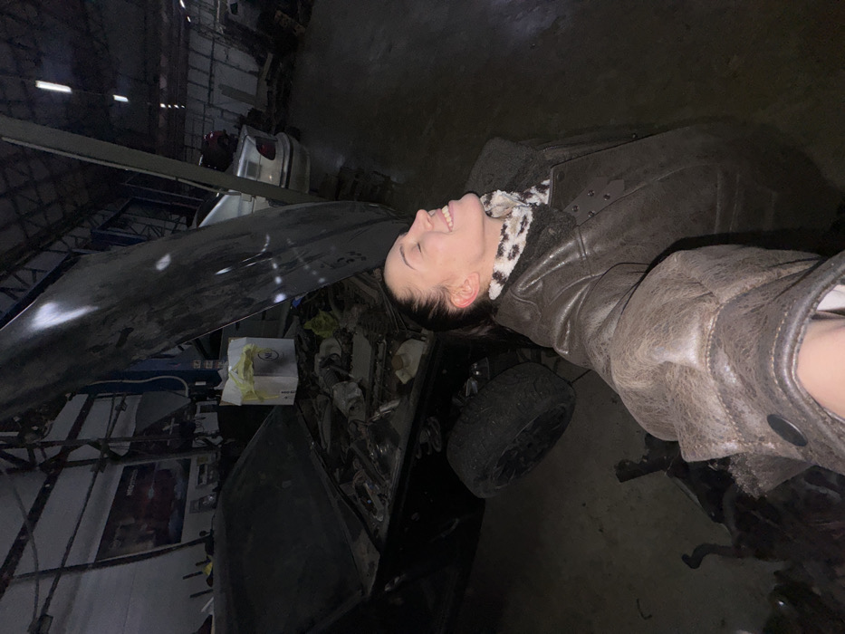 A woman in a leather jacket is laughing next to a black car in what appears to be a garage or repair shop. The car is partially disassembled, with the hood up. Tools and car parts are scattered around, and another vehicle is visible in the background. The scene is dimly lit, adding to the ambiance of a workshop setting.