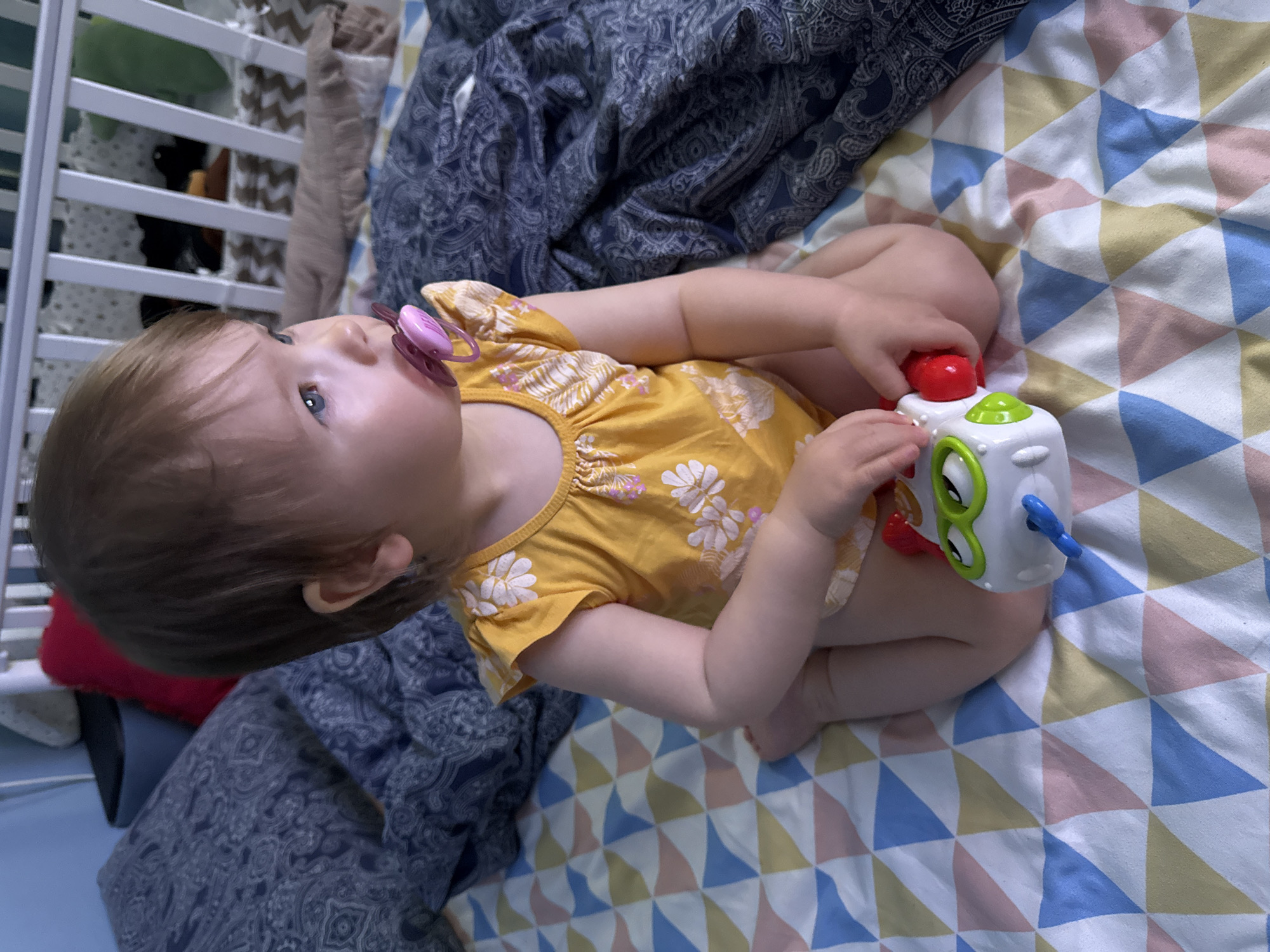 A baby with brown hair and blue eyes lies on a bed with a patterned bedsheet. The baby wears a yellow floral onesie and has a pink pacifier in their mouth. The baby is holding a white toy with green, red, and blue components. The background includes a white crib and a blue patterned blanket. It is not possible to determine the location in Moscow, Russia from the given image.