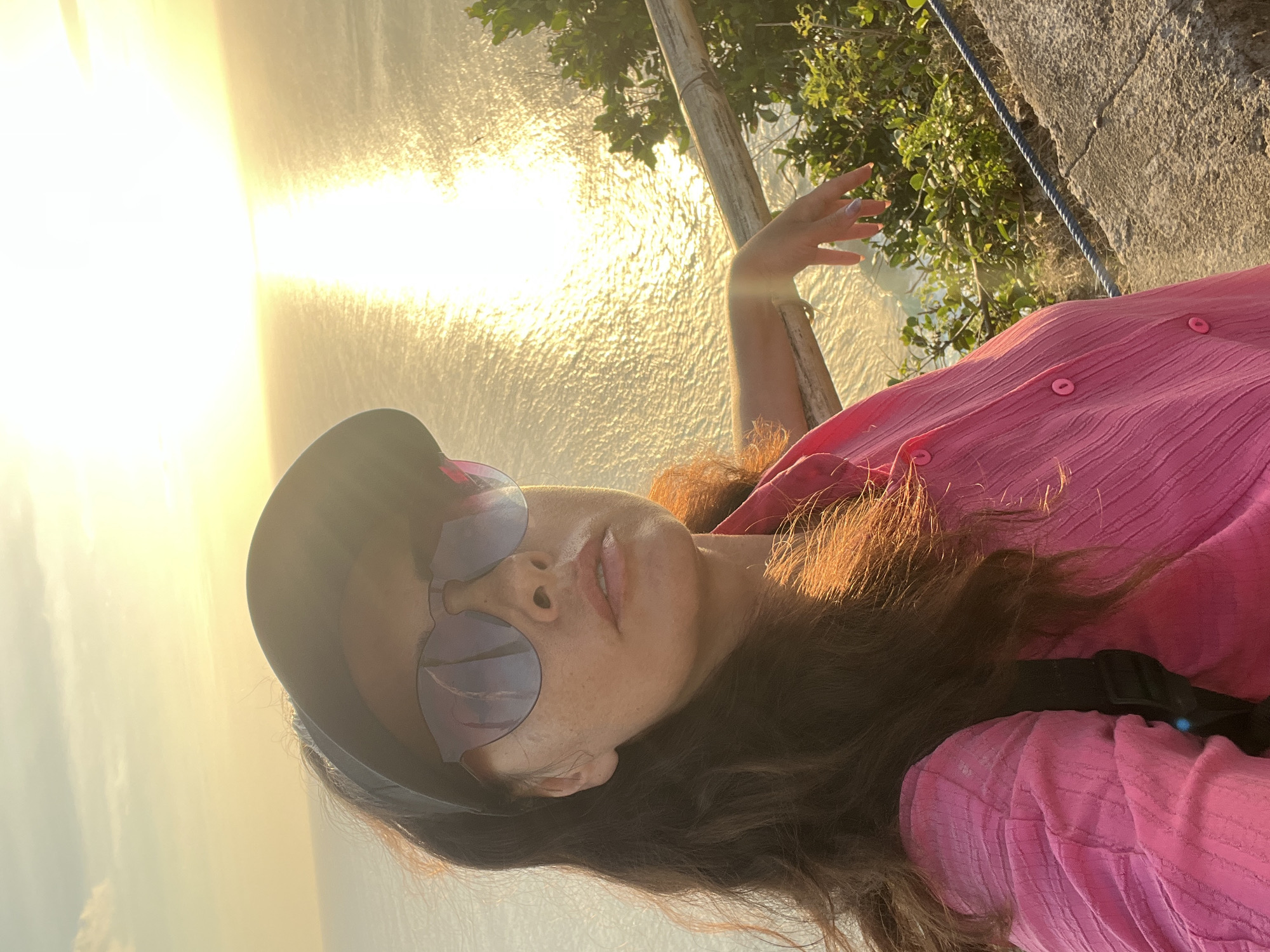 This is a close-up, horizontal, selfie-style photo taken outdoors. The subject is a person with brown hair, wearing a pink, textured button-up shirt and sunglasses. The person is leaning back and looking upwards. One hand is raised, with fingers spread, in the foreground. The background shows a body of water, possibly the ocean, reflecting the golden light of the setting sun. There are also trees and a stone wall visible. The time of day appears to be sunset due to the warm, low-angle light. The scene suggests a scenic, possibly tropical, location. The overall mood is relaxed and joyful. There is no visible text in the image.