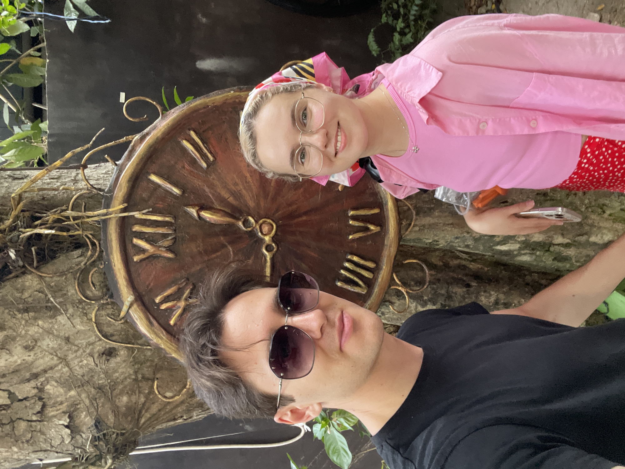 A vertical selfie captures a man and a woman positioned in front of a large, rustic clock face in an outdoor setting, likely a garden in Nam Nha Trang, Vietnam.
In the lower left, a man with dark hair wears dark sunglasses and a black t-shirt, looking towards the camera with a neutral expression. Behind him and slightly to the right, a smiling woman with blonde hair and clear-framed glasses looks directly at the camera. She is dressed in a pink t-shirt, a pink long-sleeved button-down shirt, and a red skirt with white polka dots. A colorful headscarf in shades of pink, yellow, black, and white is tied on her head. She holds a silver smartphone in her right hand, and a small flower pendant hangs from a necklace around her neck.
The prominent background feature is a large, ornate clock face crafted from wood, detailed with gold-colored Roman numerals (including I, II, IV, V, VI, VII, VIII, X, XI) and decorative hands. The clock is surrounded by golden metallic swirls and appears to be mounted on or intertwined with a textured, tree-like structure. Green foliage is visible in the background, contrasting with darker elements. The scene is brightly lit, indicating daytime.