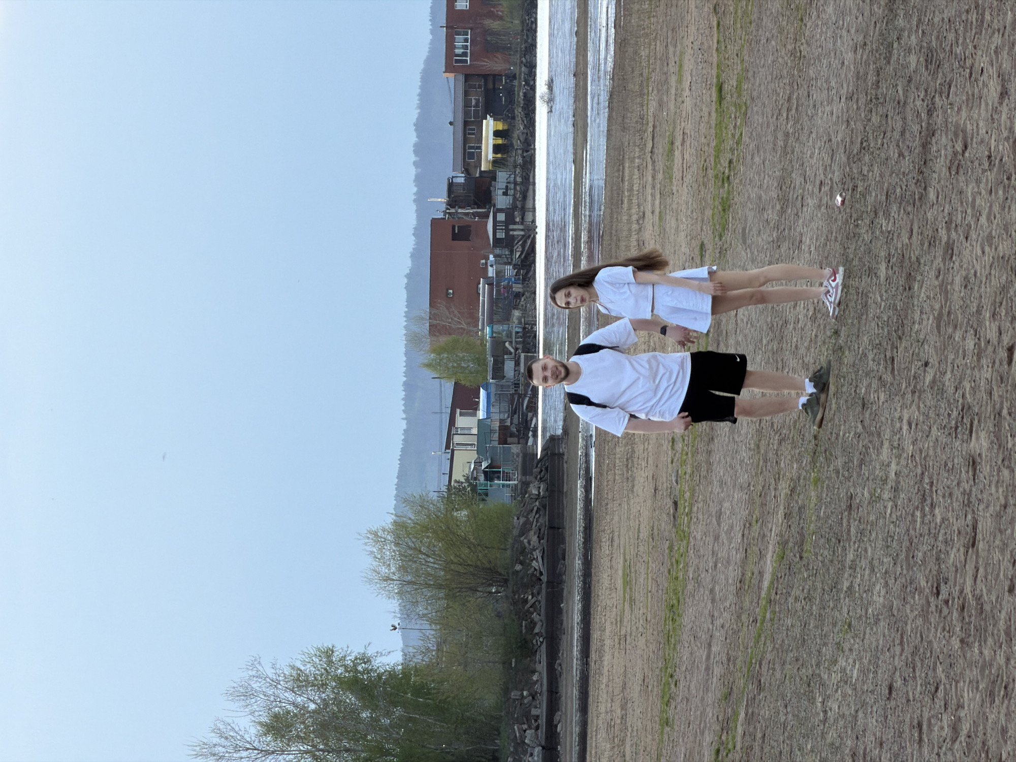 The image depicts a man and a woman standing on a sandy or muddy embankment. The man wears a white t-shirt, black shorts, and dark-colored shoes. He has a black strap, possibly from a bag, across his shoulder. The woman wears a white outfit, including a top and a skirt, and white and red athletic shoes.
In the background is the Kama River in the city of Chaikovsky, Perm Krai, Russia. Several buildings and industrial structures are visible on the opposite bank. There are trees along the near bank and in the distance, and the sky is overcast. The embankment appears to be sparsely vegetated.