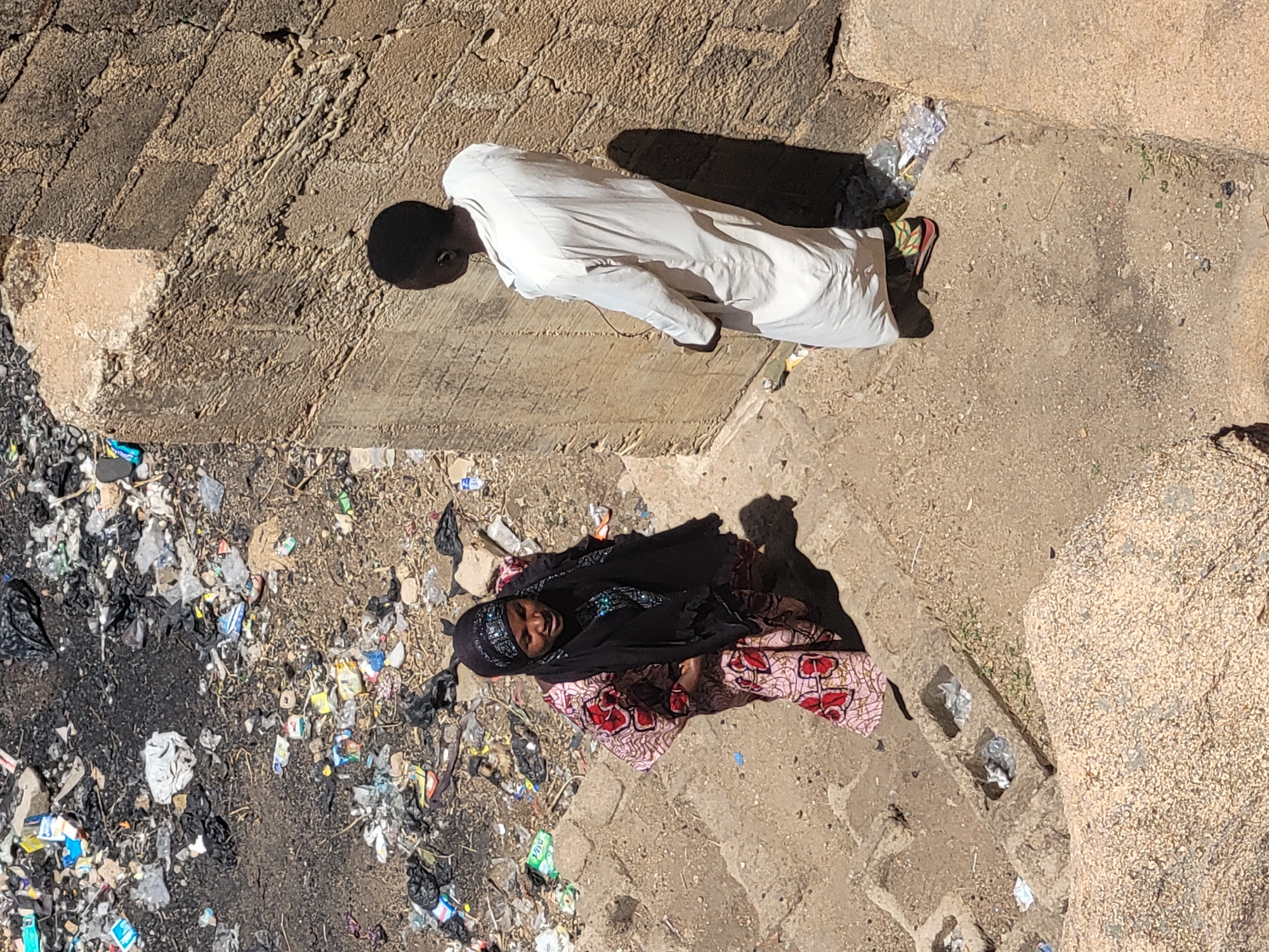 The image shows an aerial view of two individuals lying on the ground in what appears to be an urban environment. The location is Jos, Nigeria. One individual is wearing a white robe and lies on a concrete ledge. The second person is wearing a black head covering and a patterned cloth; this person lies on the ground near a large collection of trash. The ground is a mix of concrete and dirt.