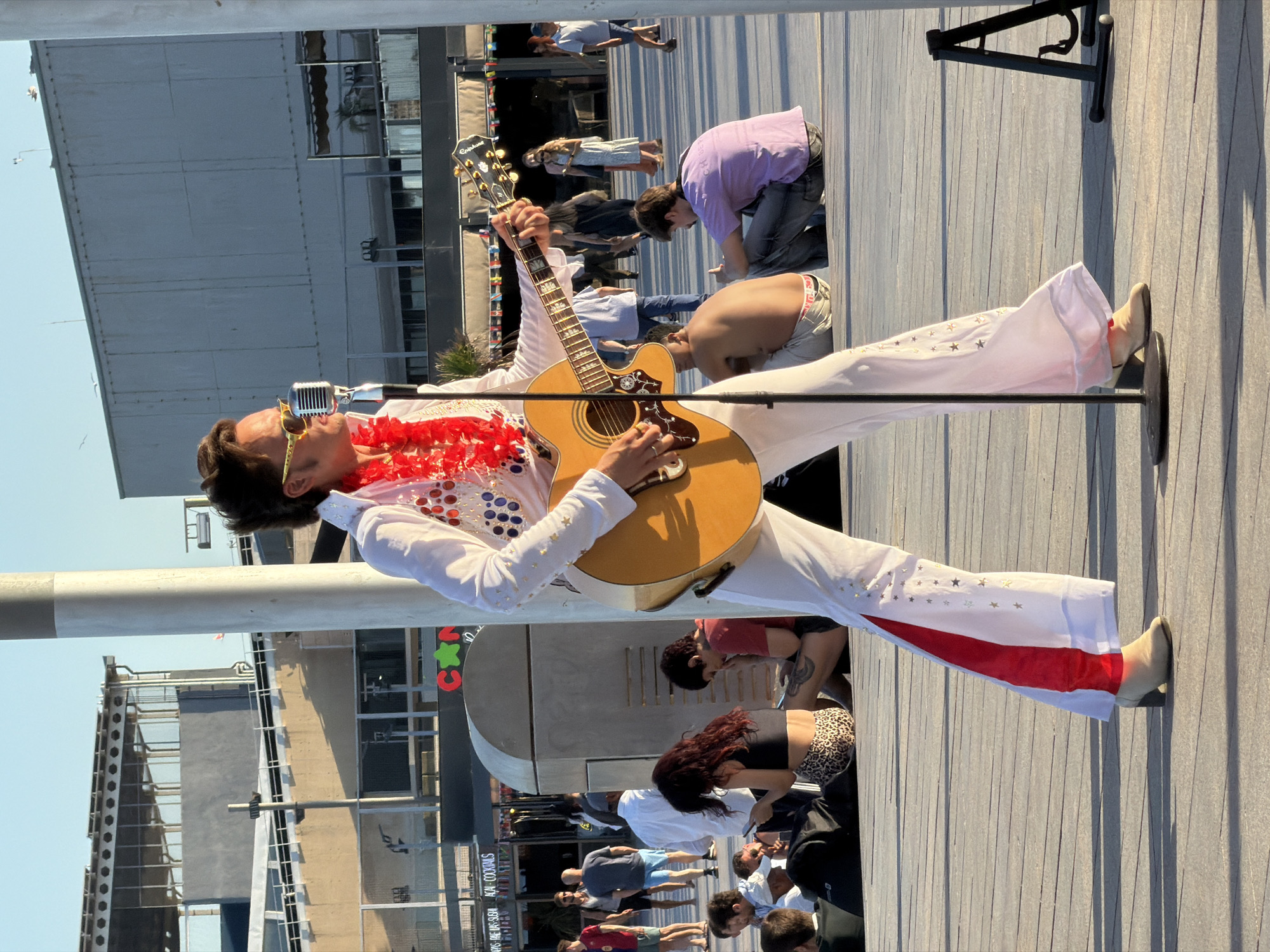 An Elvis impersonator stands on a wooden boardwalk, playing an acoustic guitar and singing into a vintage microphone. He wears a white jumpsuit with red and blue embellishments, a red lei, and yellow sunglasses. The boardwalk is outdoors, with buildings and crowds of people in the background. The scene appears to be in a public square or promenade. The photographer captured the image with a vertical orientation.