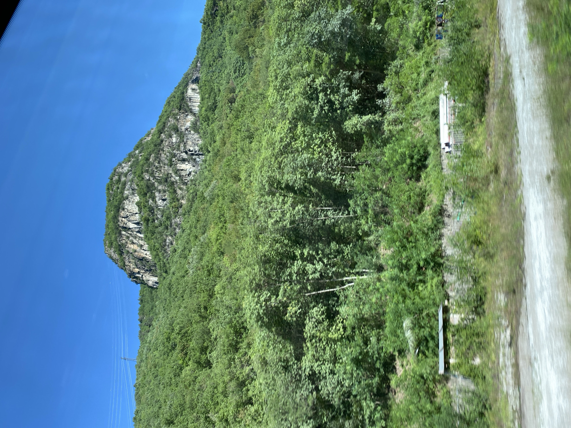 The image shows a mountainous landscape dominated by lush green trees covering the slopes. At the summit of the mountain, exposed rock faces with visible strata are present. The sky is a clear, vibrant blue. In the mid-ground, a structure resembling a small building or shelter with a corrugated roof is visible, nestled amongst the vegetation. Further to the right, along what appears to be a road or pathway, several large cylindrical objects, possibly pipes, are stacked. A few indistinct human figures can be seen near these objects. The foreground is blurred, suggesting motion and a possible perspective from a moving vehicle. The scene depicts a natural environment with some evidence of human presence and infrastructure.