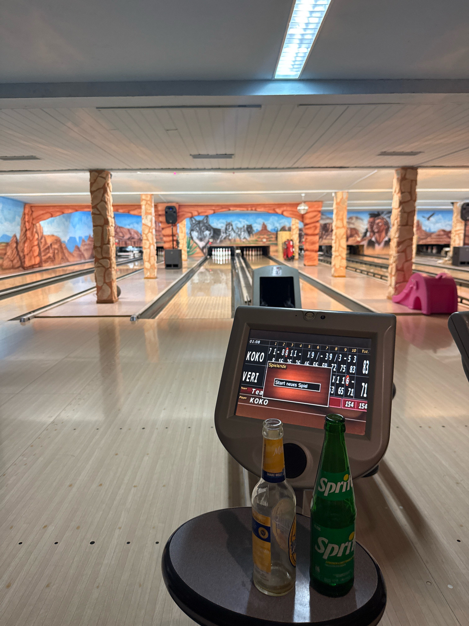 The image depicts a bowling alley with a distinct desert-themed mural on the walls, featuring rock formations and wolves. In the foreground, a bowling score monitor displays player names and scores, with "KOKO" and "VERI" listed. "KOKO" has a score of 83, and "VERI" has a score of 71. The monitor also shows a prompt to "Start neues Spiel" (Start new game). Two bottles, one empty and yellow with a blue label that reads "SHAKE WELL," and a green Sprite bottle, sit on a small table in front of the monitor. The bowling lanes stretch into the distance, illuminated by fluorescent lighting overhead. The scene is indoors, within a bowling facility in Hanau, Germany.