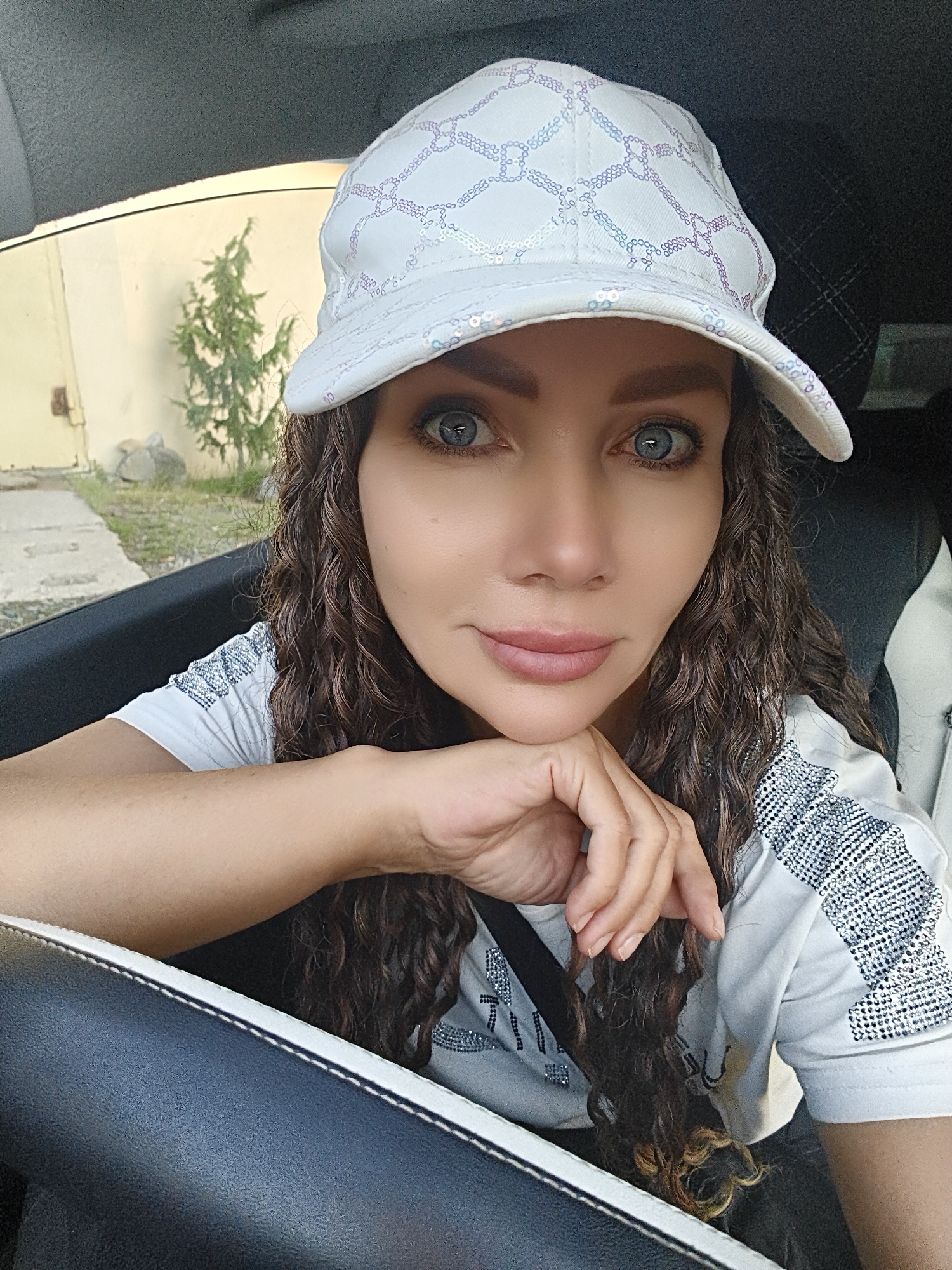 A woman with curly brown hair and blue eyes, wearing a white baseball cap and a white t-shirt with silver embellishments, rests her chin on her hand as she looks at the camera. She is sitting in the driver's seat of a car, with the car's interior visible in the foreground. The background shows a tree and a light-colored wall, suggesting an outdoor urban setting.