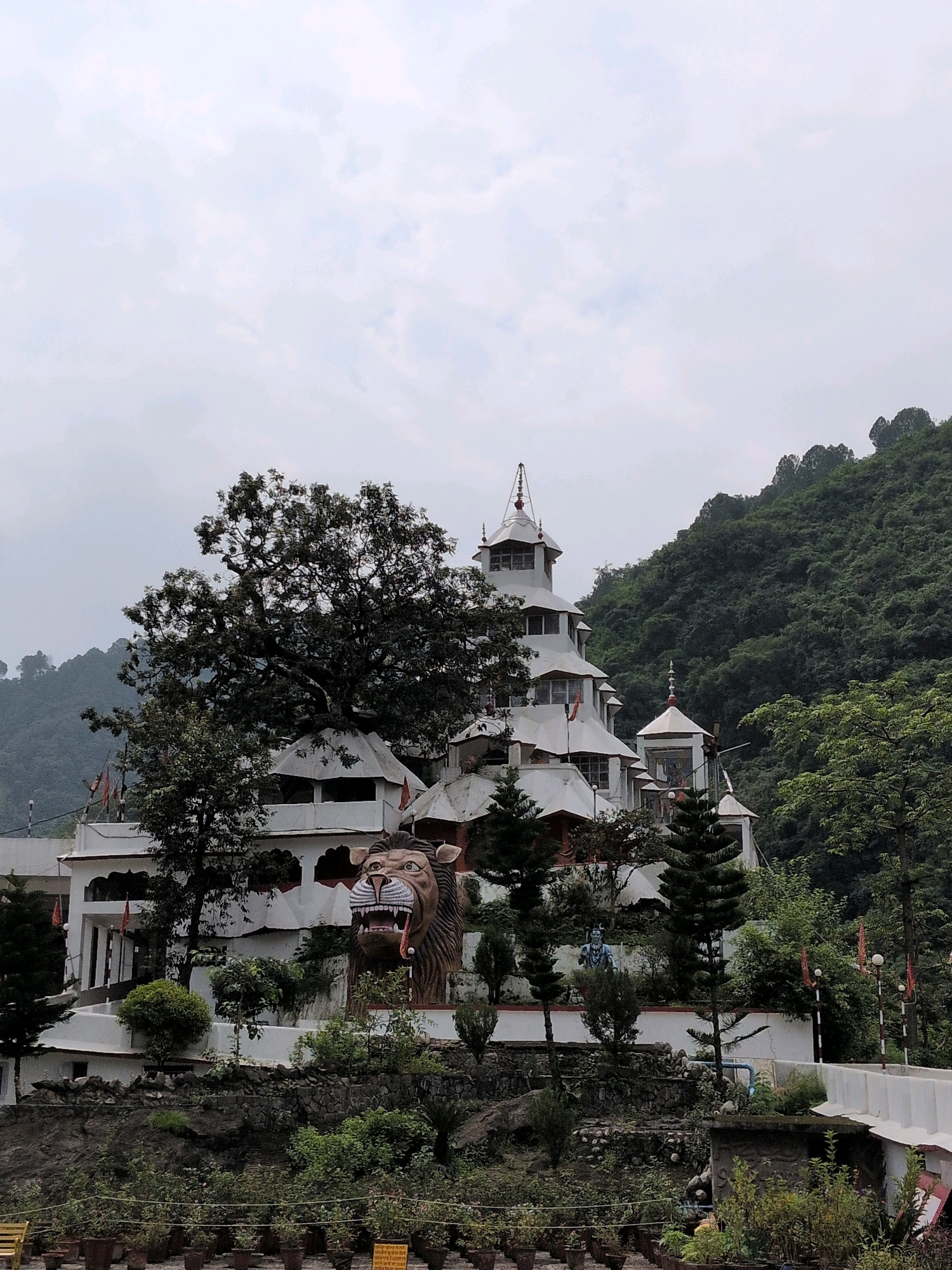 This is a photograph of the Bhimakali Temple in Sarahan, located in the state of Himachal Pradesh, India. The temple is built in a unique architectural style that blends Hindu and Buddhist influences. The structure is white with intricate wooden carvings and is surrounded by lush green hills. In the foreground, there is a large statue of a lion with its mouth open, and several potted plants are arranged along the lower terrace. The sky is overcast, and the overall atmosphere is serene and spiritual.