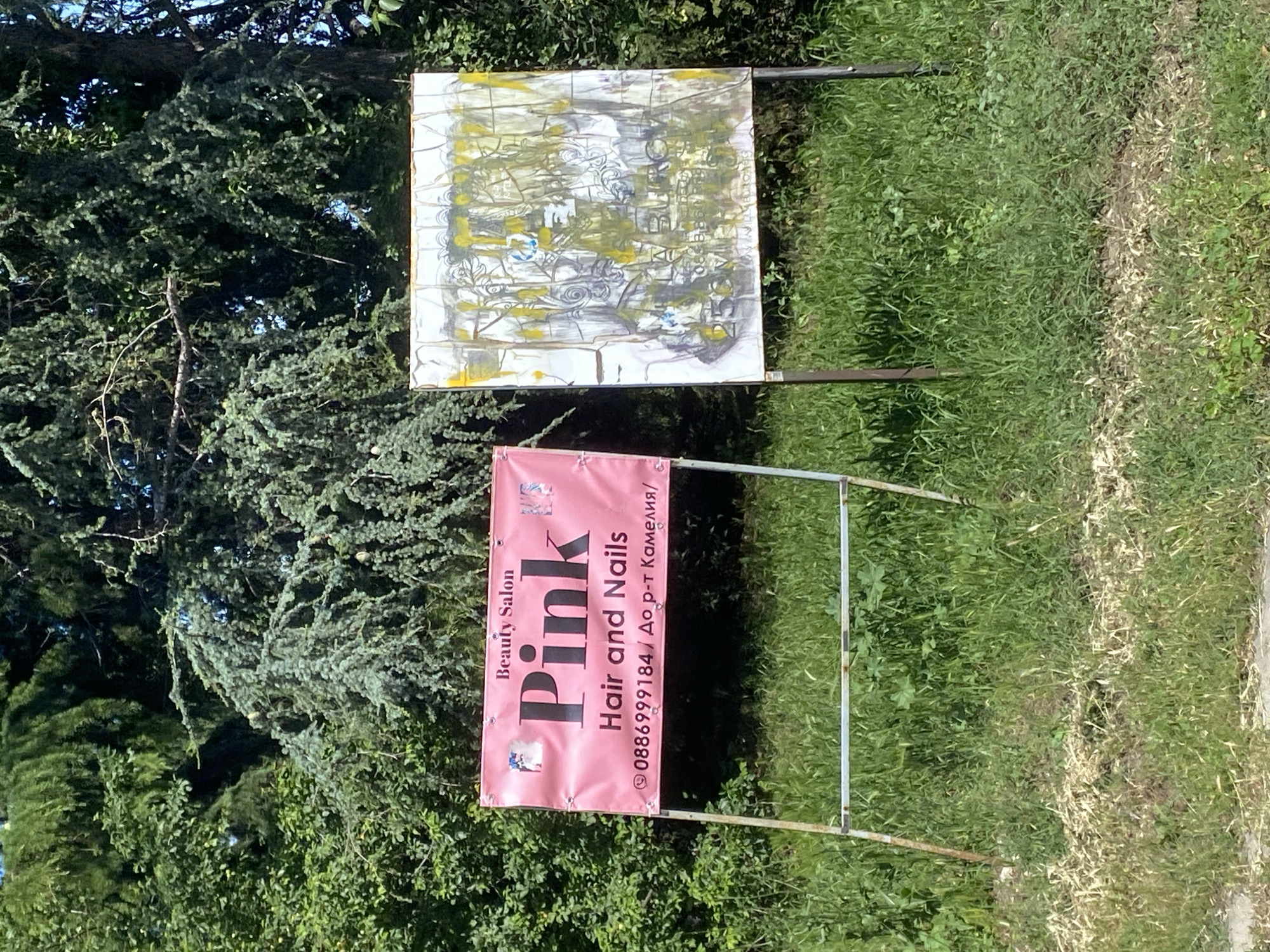 This media file shows an outdoor scene with two signs and lush green foliage. The top sign is a rectangular canvas with an abstract, textured painting featuring shades of white, grey, and yellow, possibly depicting architectural elements or a stylized landscape with swirling patterns and hints of blue. It is supported by a metal frame.
Below this, a bright pink banner displays information for a beauty salon. The text on the banner is oriented vertically and reads "Beauty Salon Pink Hair and Nails". Below this, a phone number "0886999184" and some text in Cyrillic, "До р-т Камелия/", are visible. There are also small images on the banner, possibly logos or illustrations related to beauty services. This banner is also supported by a metal frame.
The background is dominated by dense green trees and grass, suggesting a natural or park-like setting. The lighting indicates it is daytime, with sunlight casting shadows and illuminating the scene. The overall impression is of an advertisement placed in an outdoor area, likely for a local business in Varna, Bulgaria. The scene appears peaceful and ordinary.