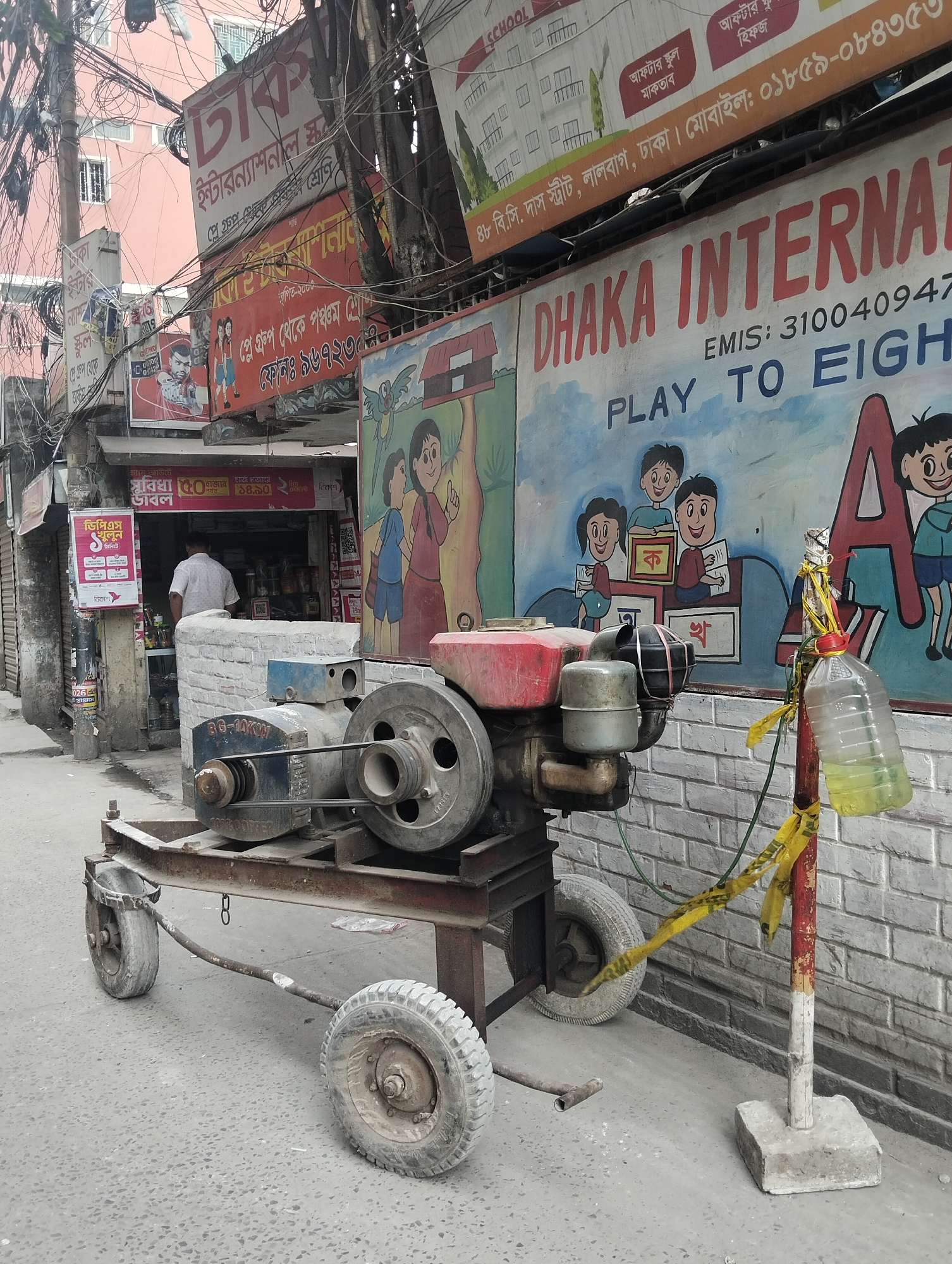 The image depicts a street scene in Dhaka, Bangladesh, characterized by a dense urban environment with an eclectic mix of infrastructure, signage, and objects.
Foreground: A portable generator sits on a wheeled frame in the foreground. A plastic bottle, possibly containing fuel, hangs from a pole nearby, secured with yellow tape.
Background: Several buildings line the street, displaying various signs and advertisements. "Dhaka International School" is visible on a prominent sign, along with other school-related information, including phone numbers and addresses. There is an advertisement on the wall. A shop can be seen in the background, with a man inside.
Setting: Overhead power lines are prevalent, contributing to the cluttered visual landscape. The overall atmosphere is that of a bustling urban street in Dhaka, with a combination of commercial and educational elements.
