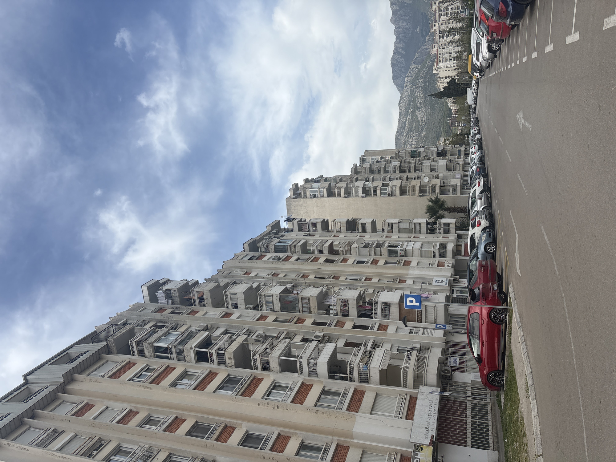 The image shows a street scene in Bar, Montenegro, dominated by a tall, aged residential building with numerous balconies. The building is off-white, with brick detailing on the lower floors. A parking sign is visible on the building's facade. A row of parked cars lines the right side of the frame, alongside an asphalt road with painted parking spaces. In the background, a mountain range can be seen under a partly cloudy sky.