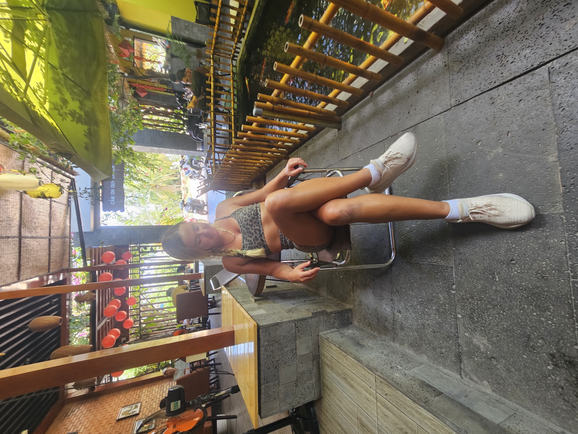 A young woman with blonde hair braided to her right side sits in a chair in what appears to be an outdoor cafe setting. She is wearing a leopard-print sports bra and matching shorts. Her legs are crossed, and she is looking down to her left, with her right hand resting on her thigh. She has on white socks and light-colored sneakers.
The cafe has a rustic, tropical ambiance. Exposed wooden beams and bamboo accents are visible throughout. In the background, a large fish pond is enclosed by a bamboo fence. Red lanterns and woven decorative items hang from the ceiling. To the left of the woman, a counter area with a wooden top is built with grey stone blocks. Further left, a large orange fan is positioned near a brick wall. Patrons are visible in the background, seated at tables, and motorcycles are parked outside. The scene is brightly lit by natural sunlight. The location is identified as Mũi Né, Vietnam.
