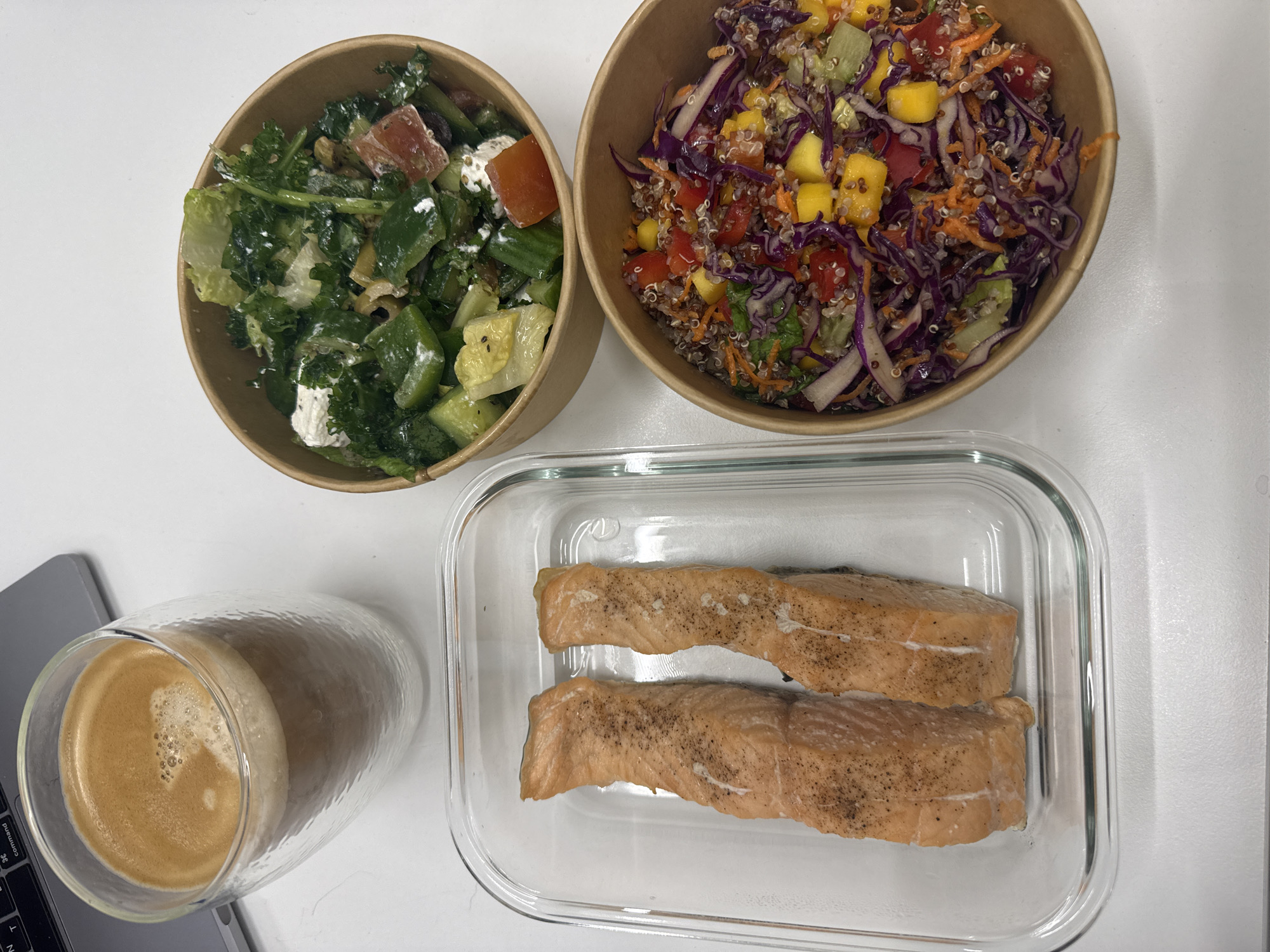 The image displays a healthy meal and a drink laid out on a clean white surface, likely a desk, next to a partially visible laptop. No people are present.
The meal consists of two salads, two cooked salmon fillets, and a coffee.
On the top left, there is a brown paper bowl containing a green salad. This salad appears to include lettuce or kale, chunks of cucumber, diced red vegetables (possibly tomatoes), and crumbles of white cheese or feta, mixed with a light dressing.
To its right, another brown paper bowl holds a colorful salad primarily composed of quinoa. This salad features shredded red cabbage, grated carrots, diced red bell peppers, and bright yellow diced mango, suggesting a vibrant and fresh mix.
Below the salads, in a clear rectangular glass container, are two pieces of cooked salmon. The salmon fillets are light orange-pink in color and lightly seasoned with black pepper.
To the left of the salmon container and partially on a gray laptop, there is a clear glass with a textured surface, holding a coffee or espresso drink with a layer of crema on top. A small portion of the laptop keyboard is visible, showing the "command" key.
The overall scene suggests a prepared lunch, possibly for an individual working from home or in an office setting. The presentation of the food indicates a focus on fresh and healthy ingredients. There are no explicit location cues related to Dubai, UAE, within the image itself.