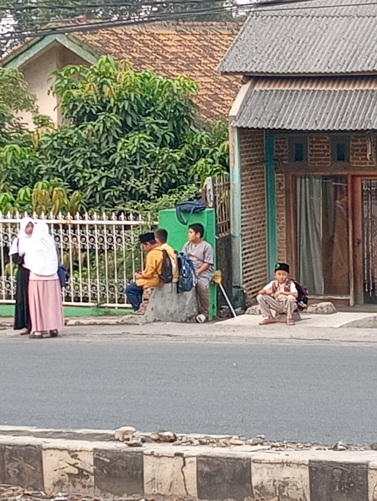 In an outdoor urban environment, likely in a residential area, two young individuals dressed in hijabs and traditional Islamic clothing stand near a white decorative fence. One is wearing a black abaya and a white headscarf, while the other is in a white hijab and a long pink skirt. Further down the sidewalk, three young males are seated on a low concrete barrier. Two of them are wearing backpacks; one is in a yellow shirt and blue pants, and the other is in a grey shirt and pants. Another young male, also wearing a backpack and a black cap, is seated on a raised concrete slab in front of a building constructed with exposed brick. The building has a corrugated metal roof and a window with green trim. Trees with lush green foliage are visible in the background. The scene appears to be a street in front of a residence or a place of worship.