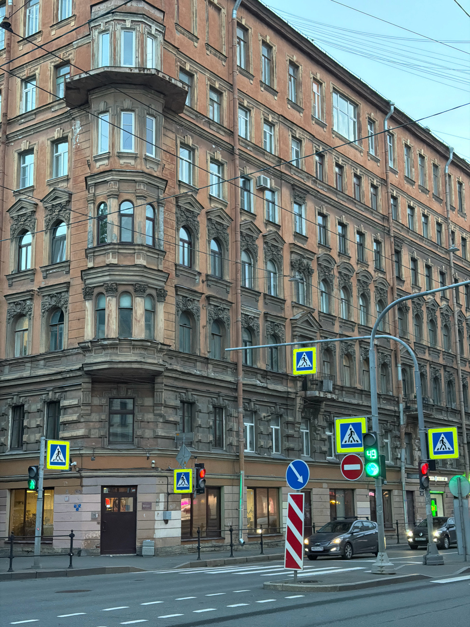 The image depicts a street corner in Saint Petersburg, Russia, featuring a prominent, ornate, multi-story building with a pale orange facade and intricate architectural details, including bay windows and decorative moldings. The building appears to be old, with some signs of wear and tear.
On the street, there are several traffic signals and road signs. A pedestrian crossing is visible, with yellow square signs indicating a pedestrian crossing area. Traffic lights show a green light on the left and a red light on the right. A digital display on the traffic light shows the number '49'. There are also circular signs with a red border, one with a white horizontal bar indicating 'no entry', and another with a white downward-pointing arrow indicating direction. A vertically striped red and white pole stands at the corner.
Two cars are present on the street. One is a dark gray sedan driving across the intersection, and another darker vehicle is partially visible behind it. The sky is overcast, suggesting a late afternoon or early morning setting. Overhead, a complex network of electrical wires crisscrosses the scene, typical of urban environments. The overall impression is a busy city intersection with a blend of historic architecture and modern traffic infrastructure.