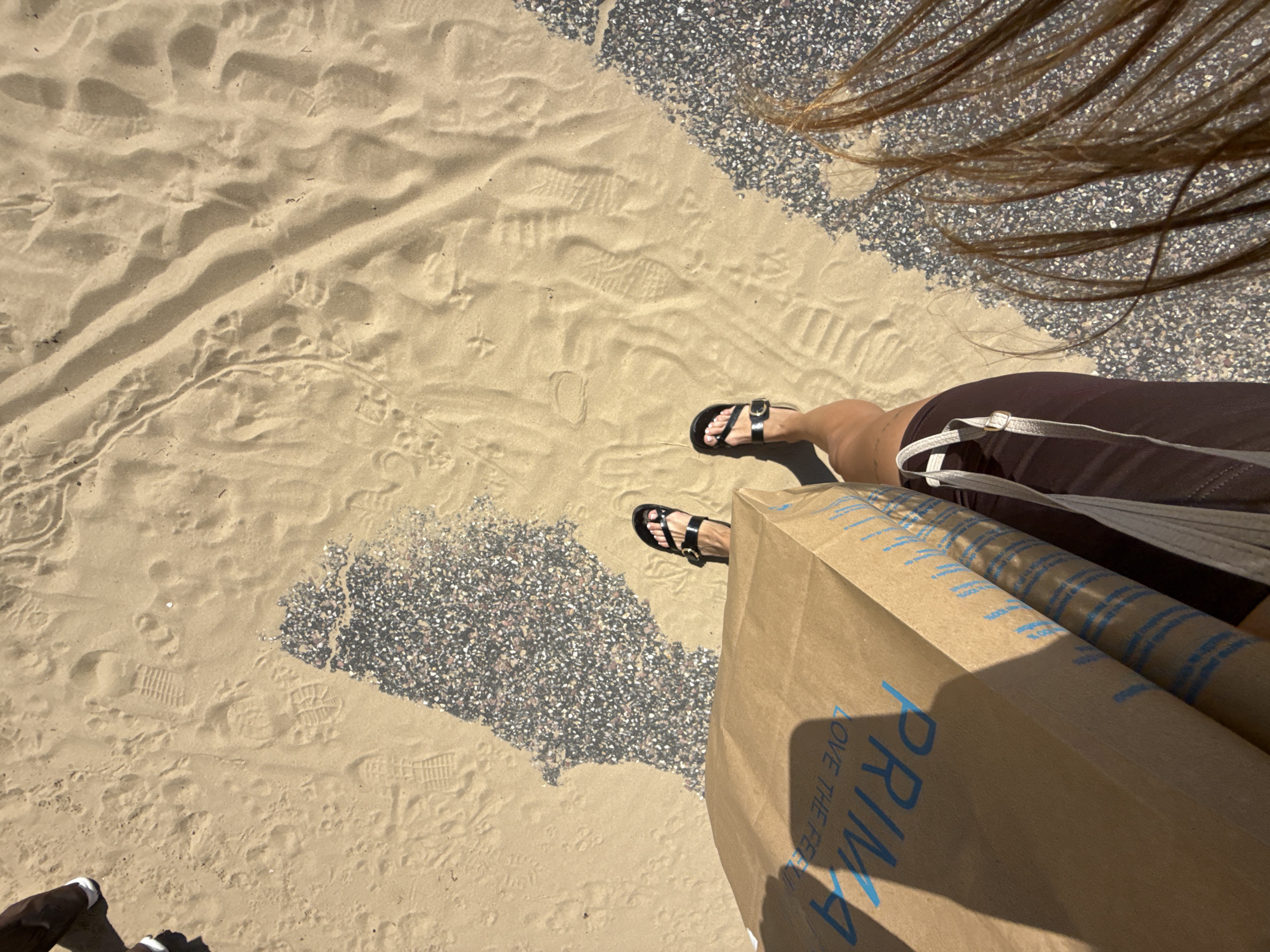The image depicts a person's legs and feet standing on a sandy area, possibly a beach or promenade, in Bournemouth, United Kingdom. The sand is marked with numerous footprints. To the right of the person, there is a section of pebbled ground. The person is wearing dark sandals and holding a brown paper shopping bag with blue lettering that appears to read "PRIMARK LOVE THE FEEL". The person's hair is long and brown and is visible in the bottom right of the frame, suggesting it is blowing in the wind. The lighting indicates it is daytime and sunny, casting shadows from the person and the shopping bag. Another person's legs and feet, wearing white sneakers, are partially visible in the upper left corner, also standing on the sandy ground. The context of Bournemouth, UK, combined with the presence of a Primark bag, suggests a shopping excursion in a coastal area.