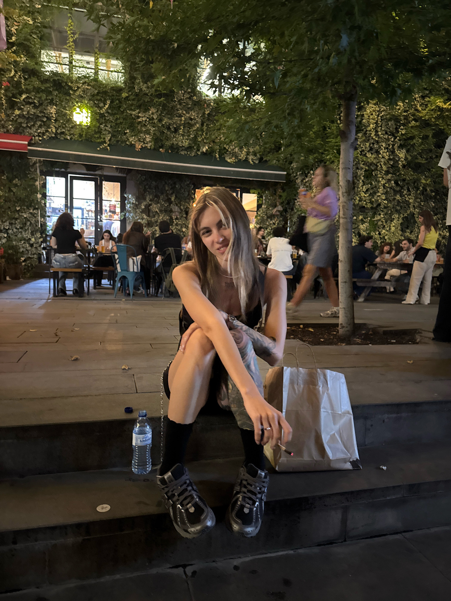 A young woman with blonde hair and bangs, wearing a black tank top, black socks, and chunky grey sneakers, sits on concrete steps at night. She has tattoos on her arms. She is holding a lit cigarette in her right hand and looking at the camera. Next to her is a brown paper bag and a bottle of water. The background shows a lively outdoor dining area with ivy-covered walls and tables with people dining. The scene appears to be in Tbilisi, Georgia, given the architectural style and vegetation.