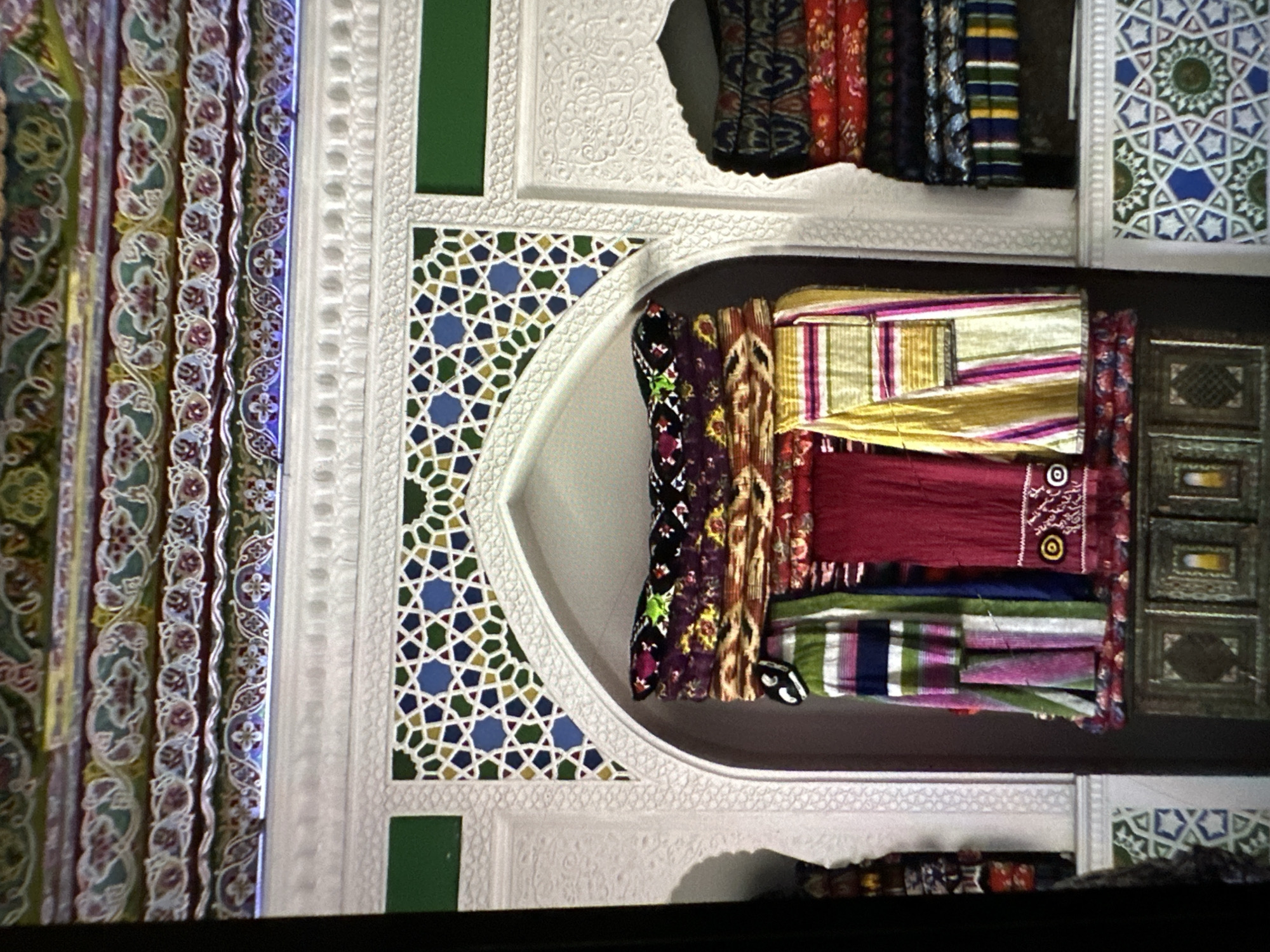 The image shows an interior view of a richly decorated space, likely a room or alcove, with vibrant textiles displayed. The architecture features intricate geometric patterns in blue, green, and yellow on white plasterwork. Above the textiles, a series of colorful, decorative friezes with floral motifs are visible along the ceiling.
In the central niche, several stacks of folded textiles are arranged. These textiles are diverse in color and pattern, with prominent examples including a dark fabric with floral embroidery, and striped fabrics in shades of green, pink, blue, and white. To the right of this central display, another alcove holds stacks of more folded textiles, predominantly in red, blue, and green with geometric and floral patterns.
The bottom portion of the image reveals a decorative wooden cabinet with carved panels and inlaid details. The overall impression is one of traditional craftsmanship and cultural richness. The location is identified as the city of Кызыл-Булак, Kyrgyzstan. There are no people visible in the image.