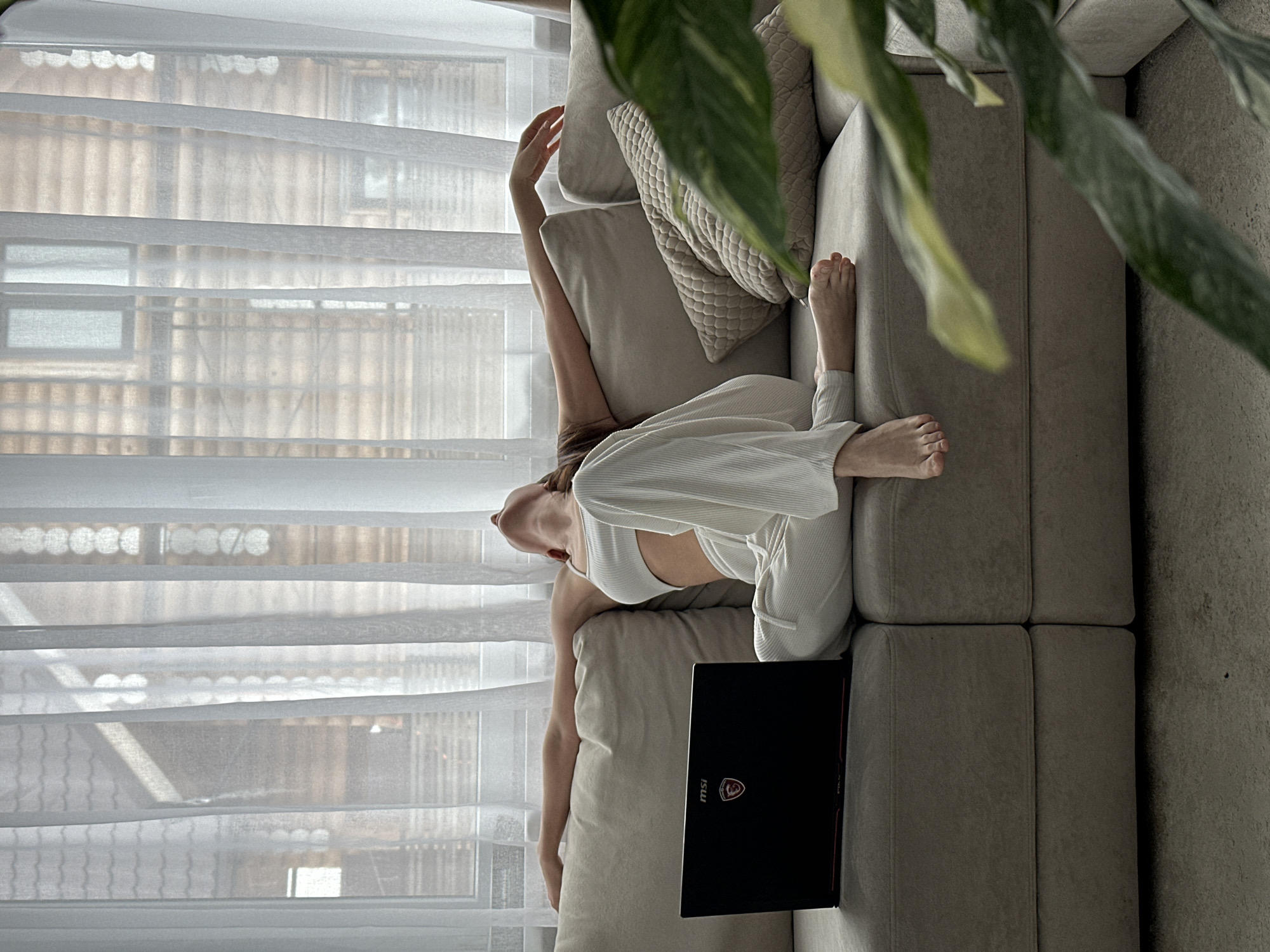 A person is lounging on a light beige sectional sofa, seemingly upside down, in a room with white, sheer curtains. The person is wearing a white top and loose-fitting pants. A black MSI laptop is beside them on the sofa cushion. A large green plant partially obscures the upper right portion of the frame. Through the curtains, there's a view of buildings. While no specific location is pinpointed in the image, I cannot determine the location in the city of Chaikovsky, Russia without more information.