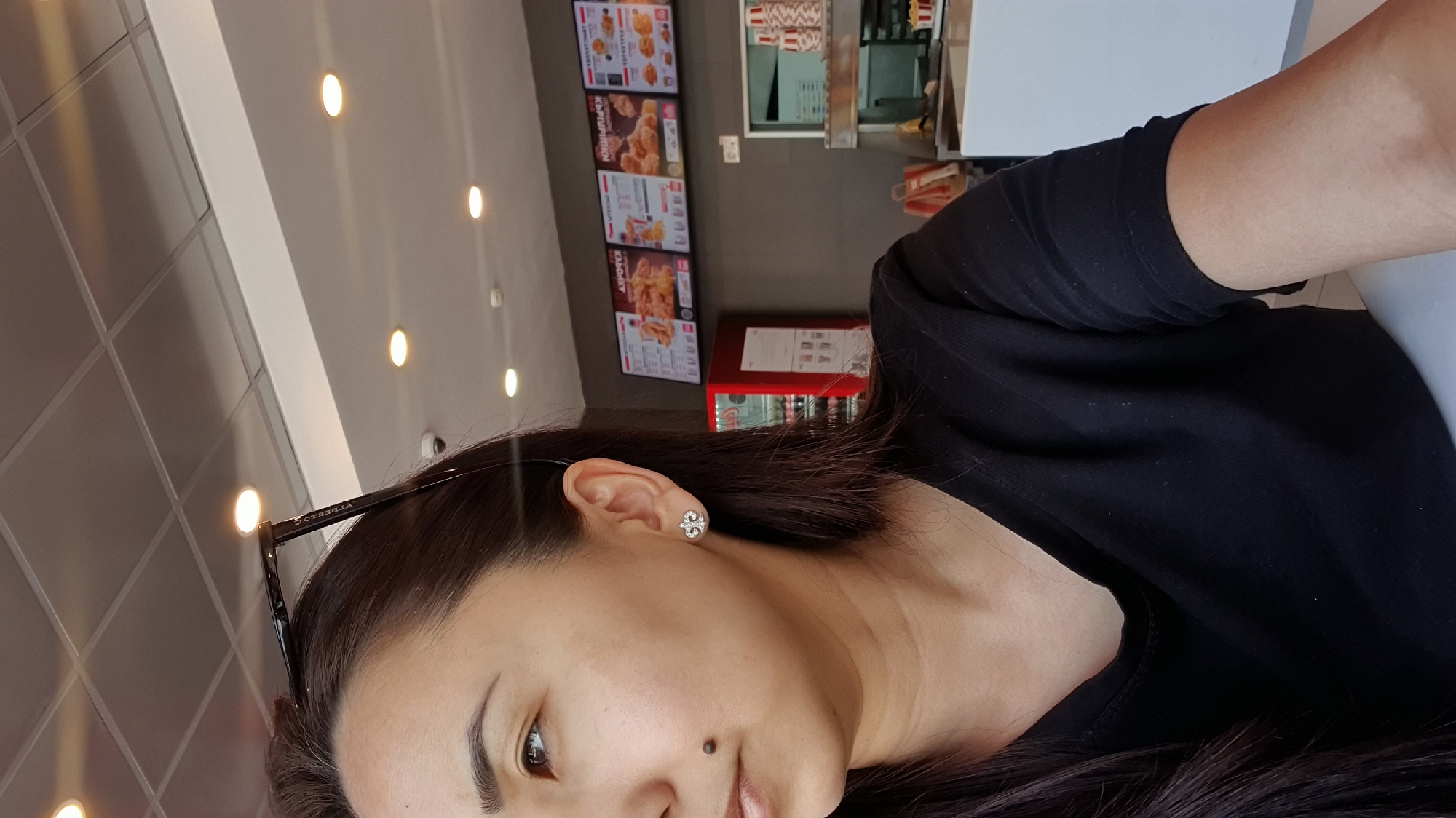 A medium shot shows a woman in a black t-shirt, seen from the chest up, looking to her left. She has dark hair and a mole on her left cheek. She is wearing a silver earring in her left ear. In the background, a fast-food restaurant is visible with menu boards displaying images of chicken meals and a red Coca-Cola refrigerator. The ceiling has square panels with embedded lights. The woman appears to be in the city administration building in Qarağandı, Kazakhstan, based on the context provided by the prompt.