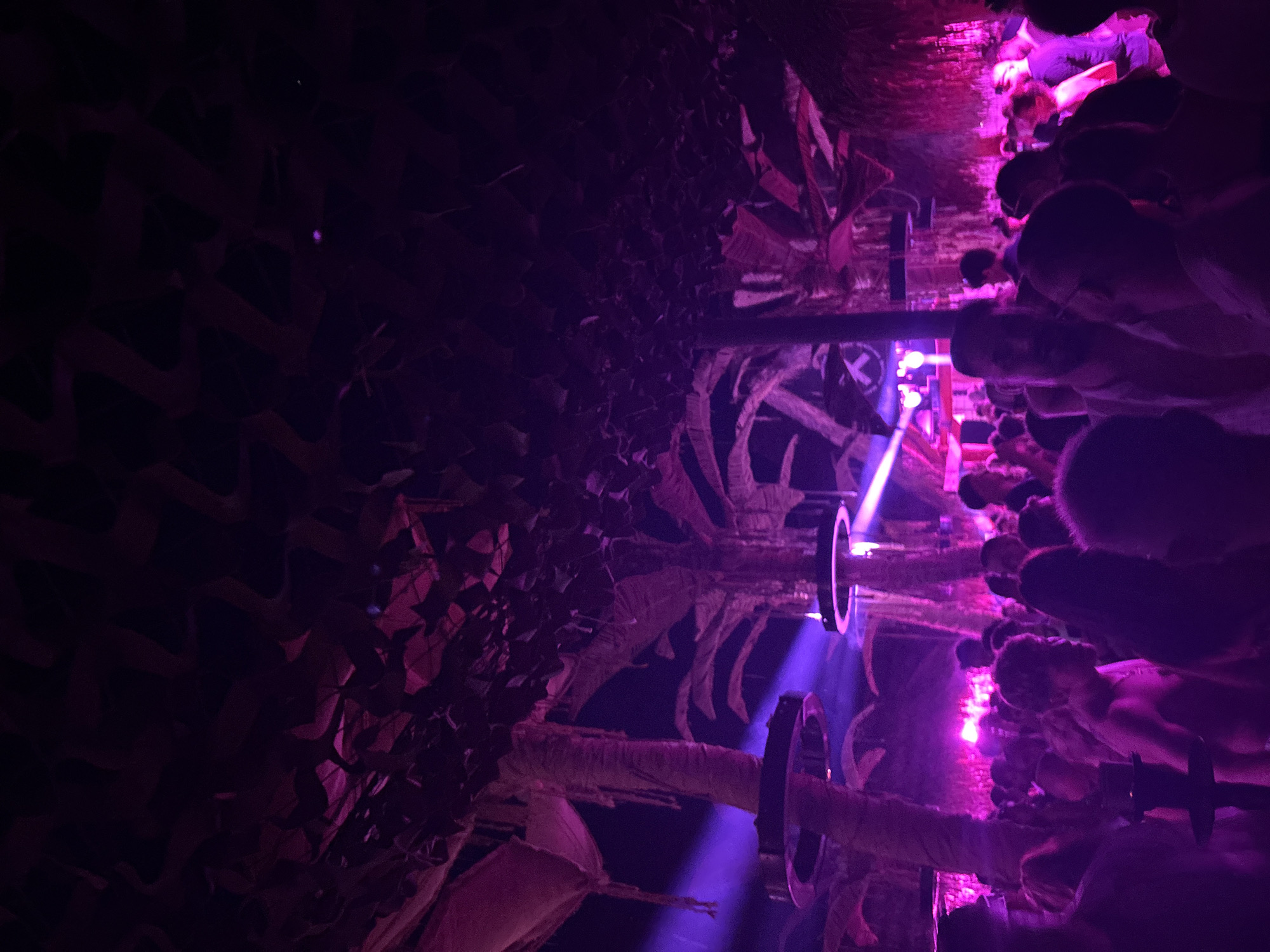 A dimly lit, crowded outdoor venue at night is depicted. The scene is bathed in purple and pink lighting, with beams of light cutting through the air. The overhead is covered with a camouflage-like netting, and some palm-like structures with hanging fabric are visible, suggesting a tropical or jungle-themed setting. A DJ booth with lights is visible in the background, where two people appear to be performing. A large crowd of people are standing and socializing. The overall atmosphere is energetic and festive, indicative of a nightclub or party.