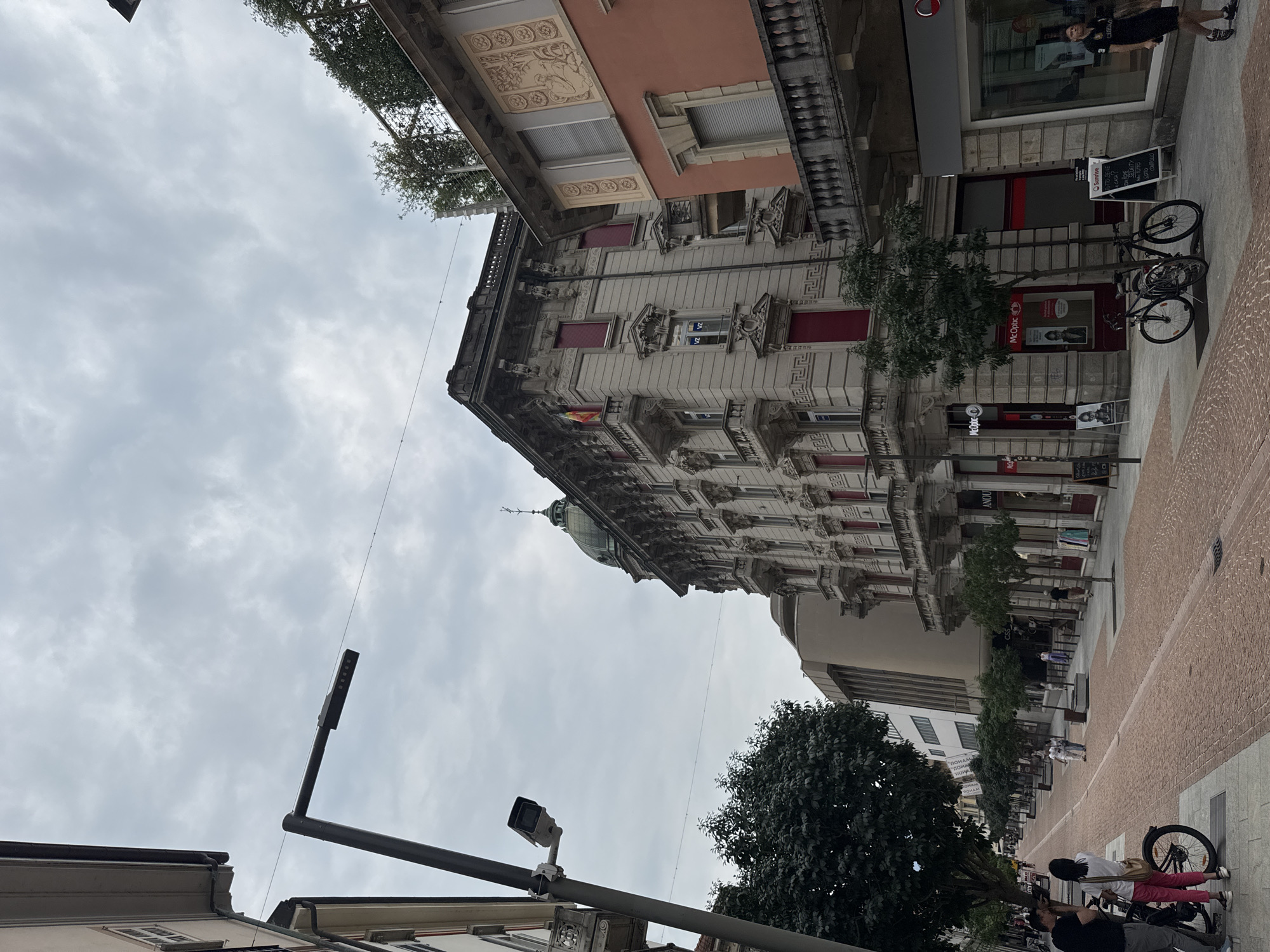 The image depicts a street scene in Bellinzona, Switzerland, on a cloudy day. In the foreground, a pedestrian walkway paved with reddish-brown bricks is visible, with a darker grey sidewalk on the left. Several people are present. On the left, a man and a woman stand next to a bicycle. Further down the street, two individuals are walking away from the camera. On the right side of the street, two bicycles are parked near a sidewalk.
The architecture of the buildings lining the street is varied. On the left, a modern building with a glass facade is visible. To its right, a grand, ornate building with a dome and balconies dominates the mid-ground. This building features classical architectural elements, including columns, decorative carvings, and red-framed windows. On the far right, another building displays a fresco-like artwork above its windows and has a stone balcony.
Various businesses are identifiable on the ground floors of the buildings, including "McOptic" and "Sunrise." Streetlights and trees are also present, contributing to the urban streetscape. The overall impression is of a busy, well-maintained urban environment.