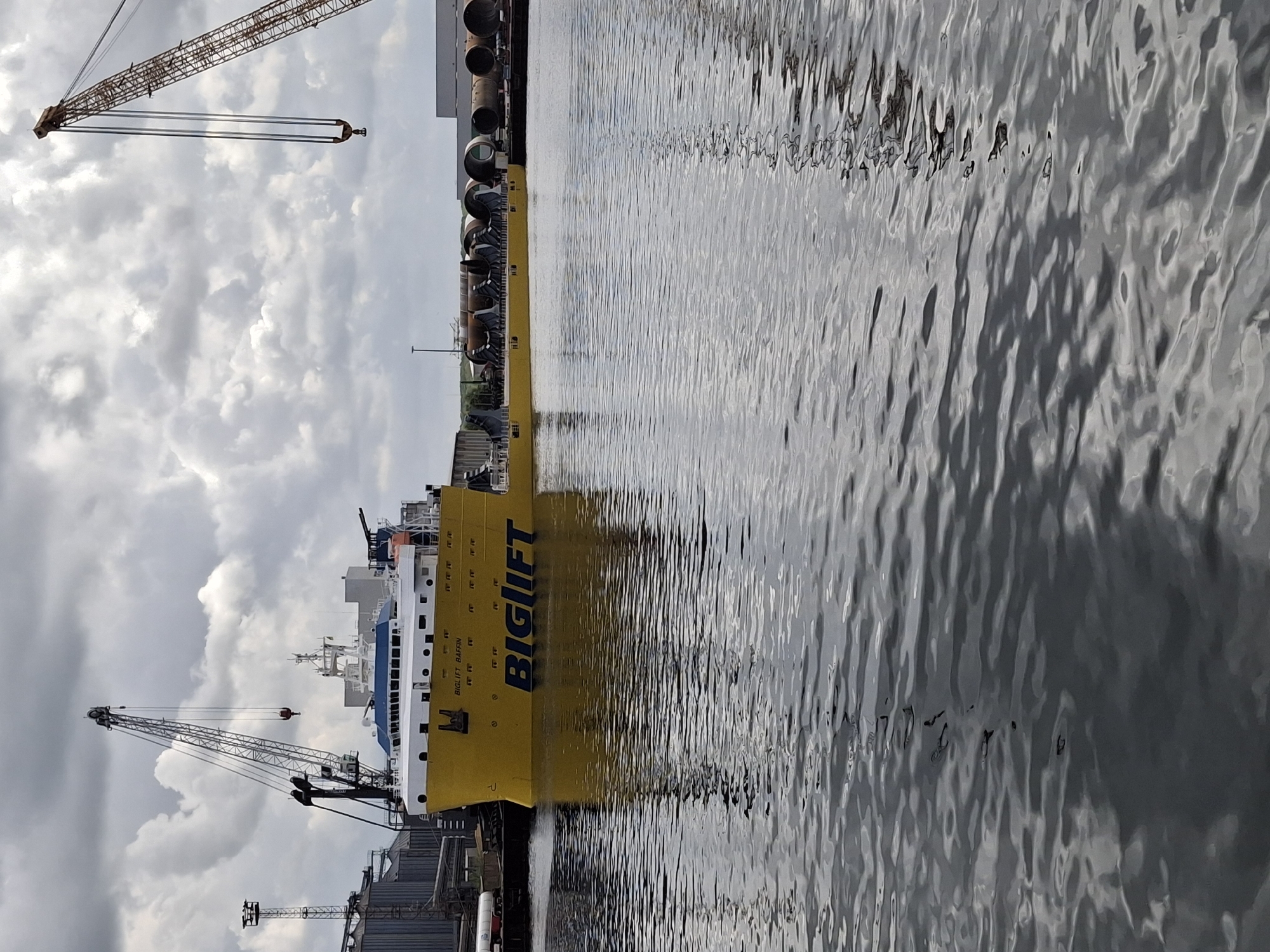 The image shows a yellow and white BigLift Baffin ship docked in Rostock, Germany. The ship is in the foreground, with the water of the port filling the right side of the frame. Various pipes are stacked on the deck of the ship. Cranes are visible near the ship and in the background, with a cloudy sky above. Dockside structures are seen at the base of the ship near the stern.