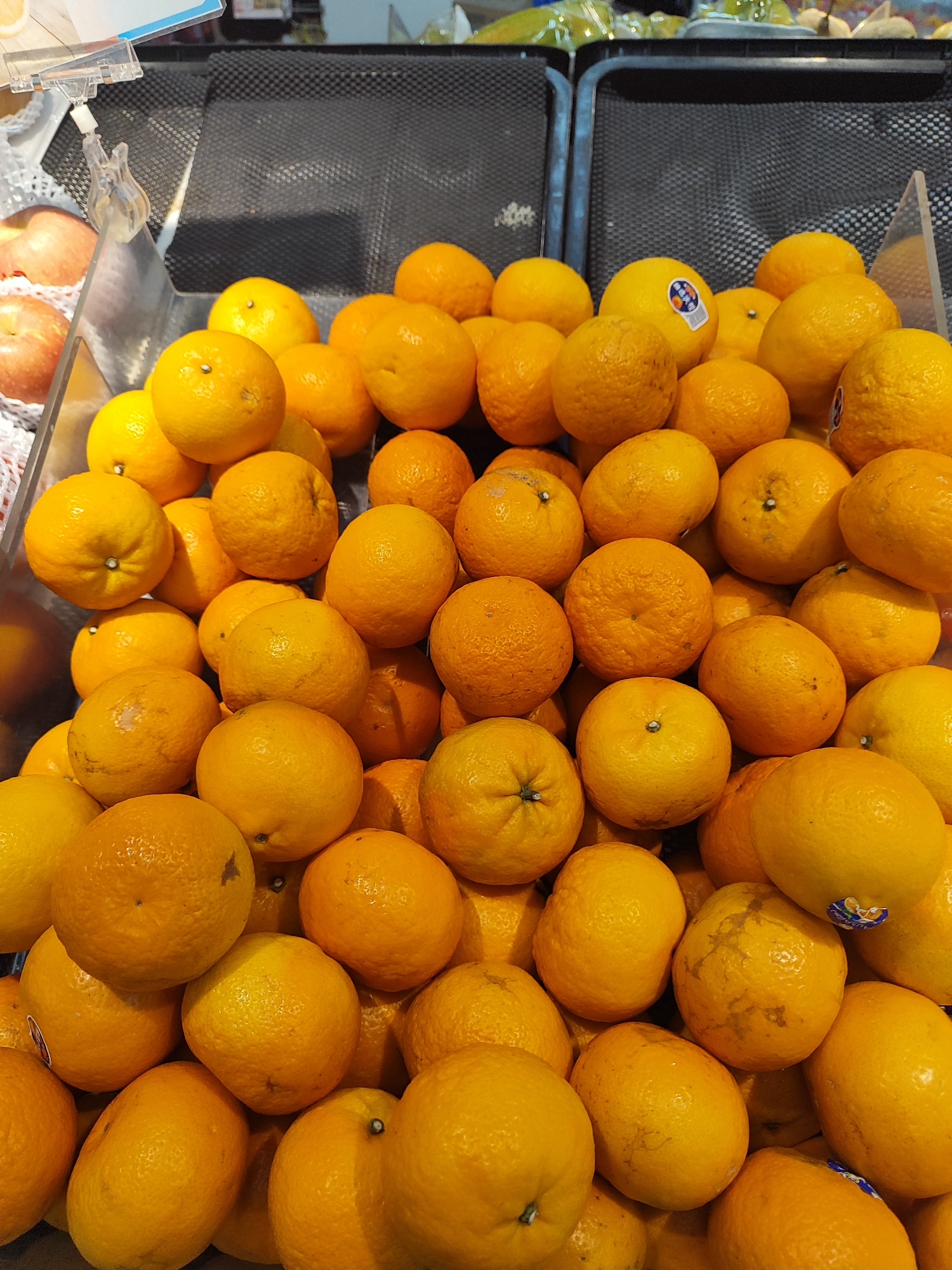 A close-up, eye-level shot shows a large pile of oranges in a supermarket display. The oranges are bright orange and vary in size and ripeness. Some have stickers on them. The background is blurred, showing black plastic bins and some green produce, suggesting a grocery store setting. The lighting is bright, highlighting the textures of the orange peels. The oranges are likely intended for sale in Nangkaan, Indonesia.