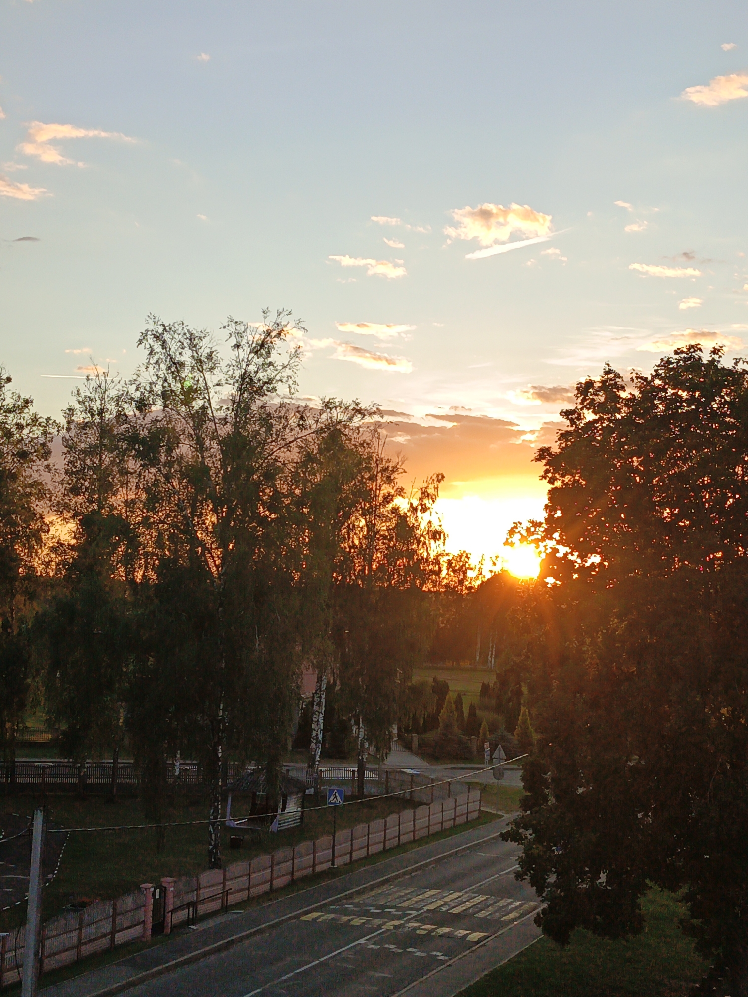 The image depicts a sunset view in the city of Parechcha, Belarus. The sky is a gradient of pale blue to soft orange, with scattered white and pink clouds. A bright sun is setting behind a dense line of trees, casting long shadows and creating a silhouette effect. The sun's rays are visible, fanning out through the foliage.
In the foreground, a road runs horizontally across the image. It has yellow and white crosswalk markings. A light-colored wooden fence runs along the side of the road, with a smaller pink-topped fence in front of it on the left. Beyond this fence, there are grass and more trees. Further back, a row of shorter trees and bushes are visible, and behind them, a longer wooden fence. A small, rustic wooden structure with a pointed roof and an open side is located in the middle ground, near the longer fence. A blue triangular sign with a pedestrian symbol is posted to the right of this structure. The overall scene suggests a quiet, possibly rural or suburban area during a picturesque sunset. There are no people or animals visible in the frame.