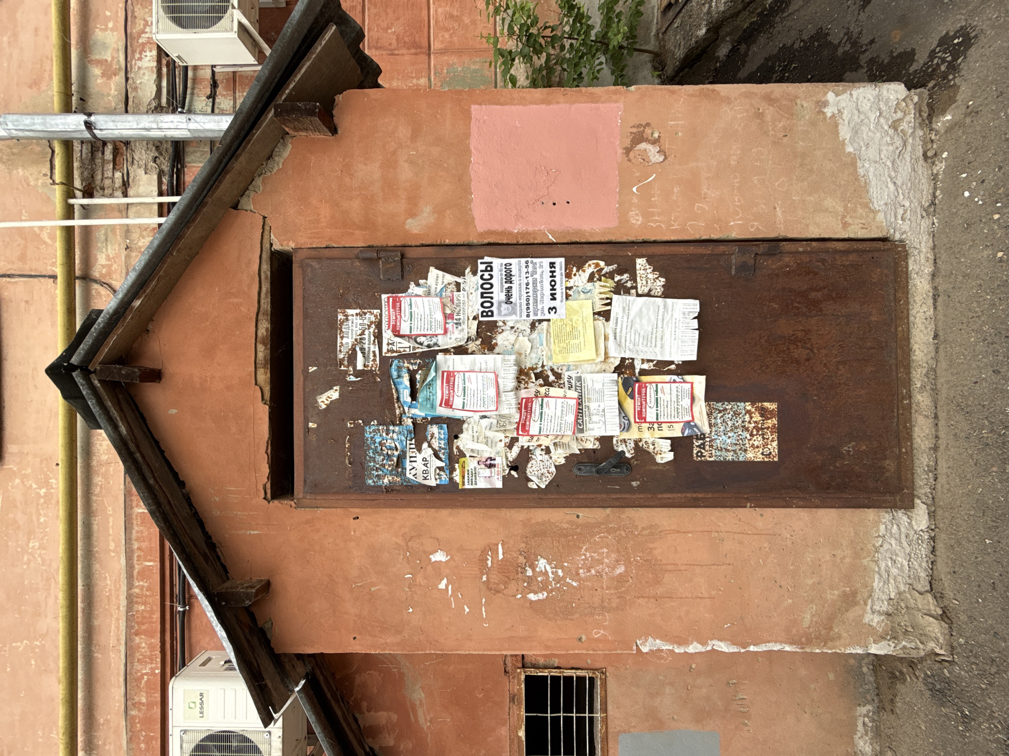 The image shows the exterior of a building in Votkinsk, Russia. The facade is primarily pinkish-orange and appears worn, with patches of discoloration and repair work visible. A rusted metal door is flush with the wall, covered in numerous tattered flyers and posters, some advertising "HAIR - very expensive, 8(950)176-13-56 hairdressing salon 'Megsi' st. Koroleva, 21, 3 June" and "BUY APARTMENTS" There is a small, barred window below the door. Above the door, a square of the wall has been painted a slightly different shade of pink. A slanted wooden structure, possibly part of a roof or awning, juts out from the upper left corner of the frame. An air conditioning unit with the brand "LESSAR" is also visible on the upper left of the frame. A section of the sidewalk and possible street is also present.