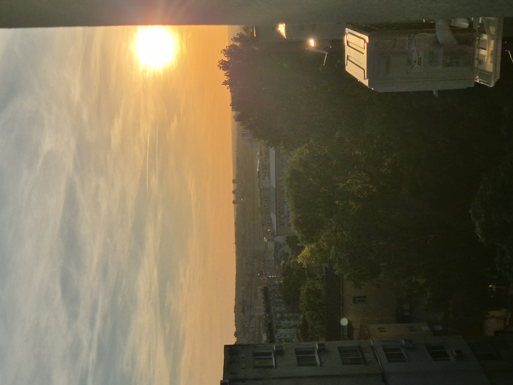 The image shows a sunset over a cityscape. The sun is prominently visible, casting a warm golden light across the sky and cityscape. In the foreground, there's a building with several windows. A tree line is present with buildings interspersed in between. There also appears to be an object that is reflecting the light, possible a window or other reflective structure. The city extends into the distance.