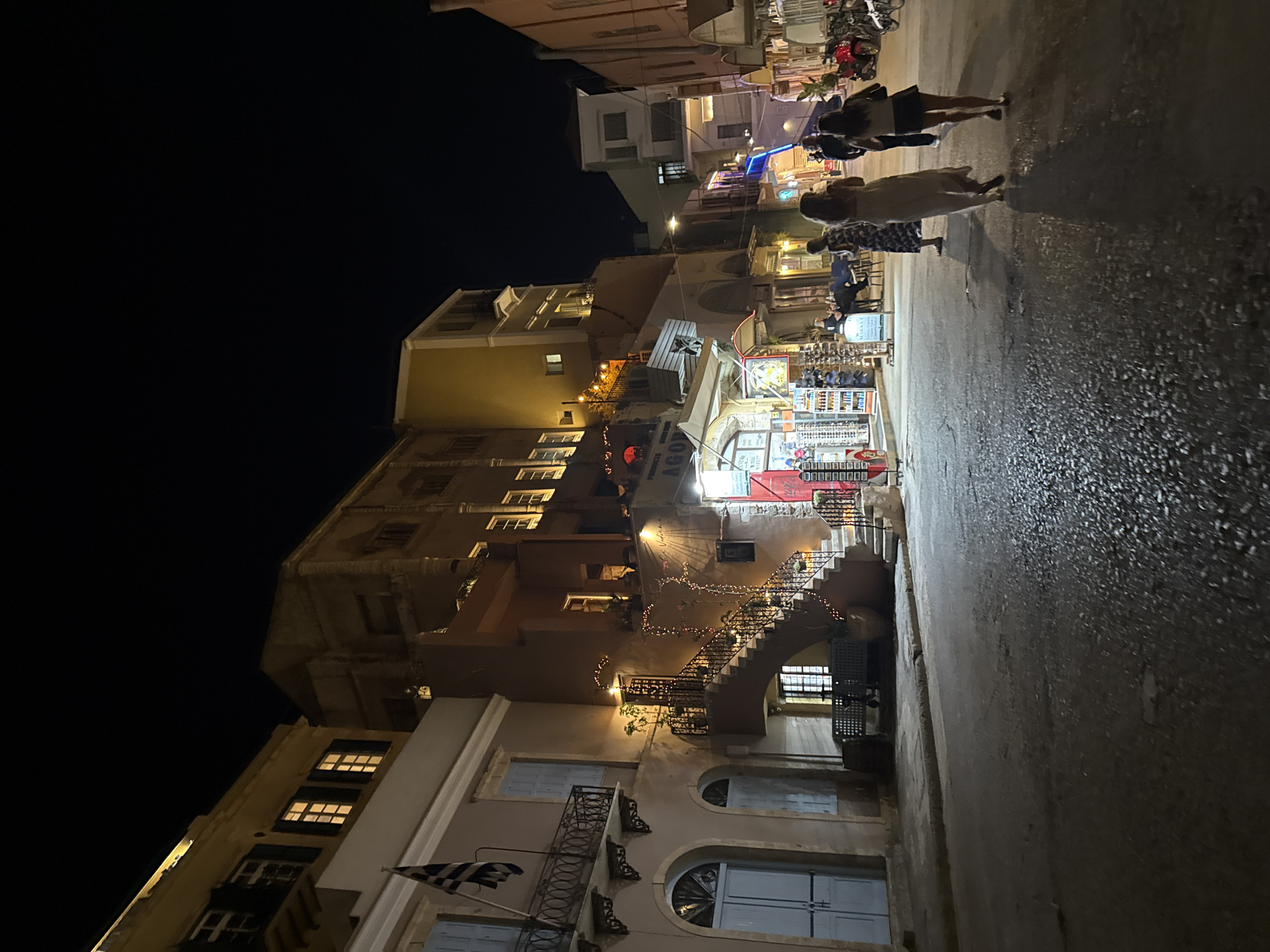 The image depicts a nighttime street scene in a European city, likely in Greece given the flag visible on the left. The street is paved with cobblestones and appears wet, reflecting the ambient light from shops and buildings. On the left, a pale pink building features arched doorways and balconies. To the right of this building, a multi-story structure in a warm, earthy tone has an external staircase adorned with fairy lights leading up to an entrance. Shops are open along the street, with illuminated displays of merchandise, including what appears to be souvenirs and clothing. Several individuals are walking along the street, some in groups, suggesting a lively evening atmosphere. In the background, the street continues, lined with more buildings and shops, and a few parked motorcycles are visible. The sky is dark, indicating it is night.