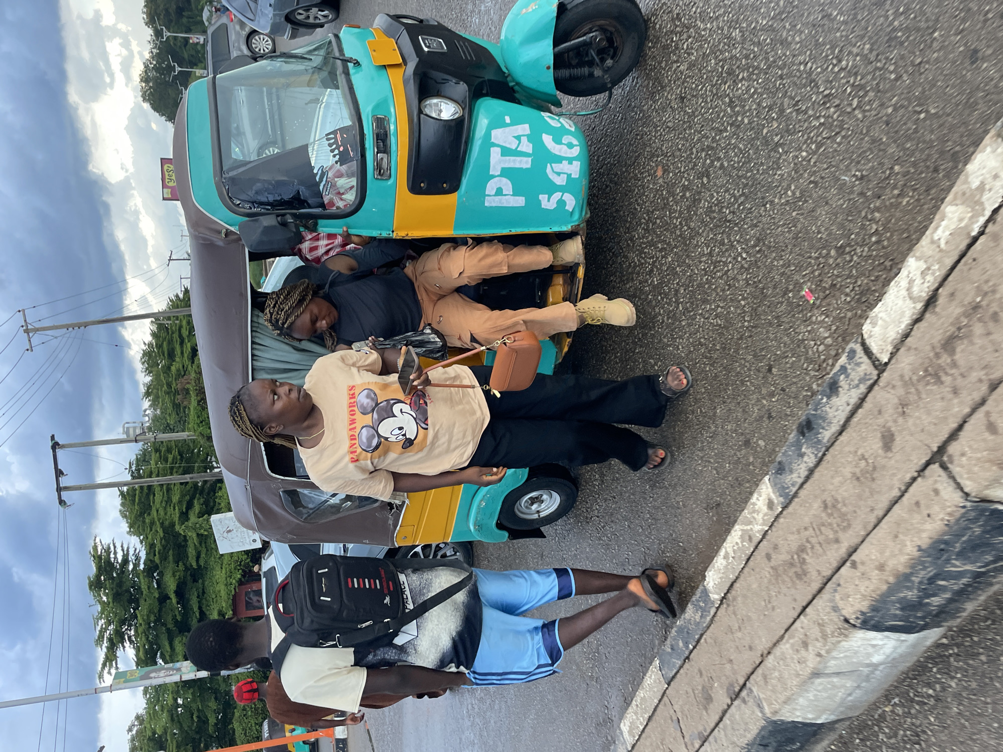 A low-angle shot captures a turquoise and yellow auto-rickshaw with three people interacting around it on a wet asphalt road in Jos, Nigeria. The auto-rickshaw has the license plate "PTA 5462". A man wearing a tan t-shirt, blue shorts, and black flip-flops with a black backpack walks away from the auto-rickshaw. A woman with braided hair, wearing a tan t-shirt with a Mickey Mouse graphic and black pants, stands near the open door of the auto-rickshaw, holding a mobile phone in her right hand and a brown purse in her left. Another woman with braided hair is seated inside the auto-rickshaw, wearing a black top and tan cargo pants. The environment includes a concrete curb with black and white markings, traffic lights, power lines, and trees in the background. The sky is cloudy.