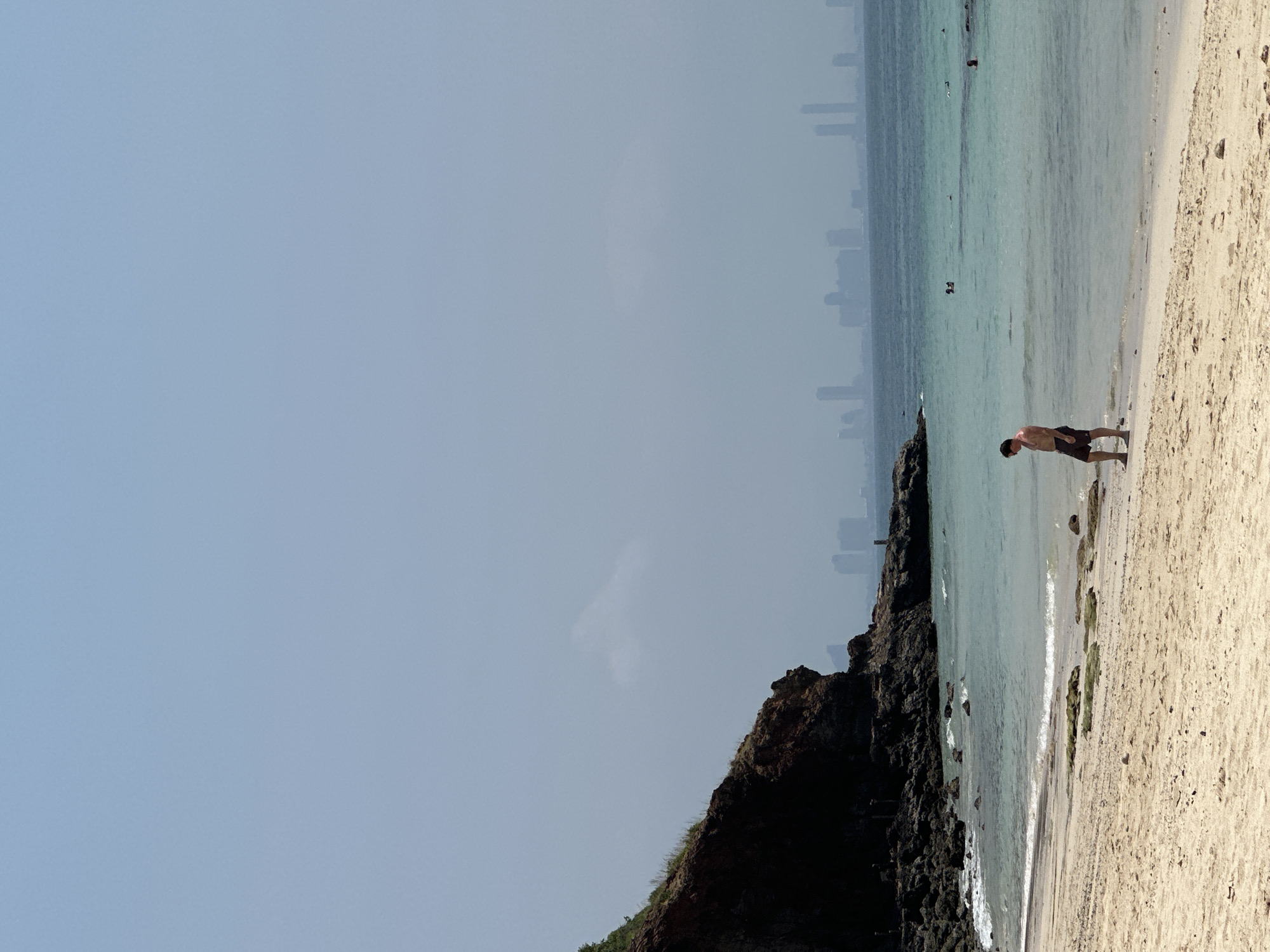 The image shows a beach scene with a man standing near the shoreline. A rocky cliff borders the left side of the beach. The water is a clear turquoise color. In the distance, a faint skyline of Pattaya City, Thailand, is visible. Other people are present in the water further out. The sand is light in color, and there are some small rocks near the waterline.