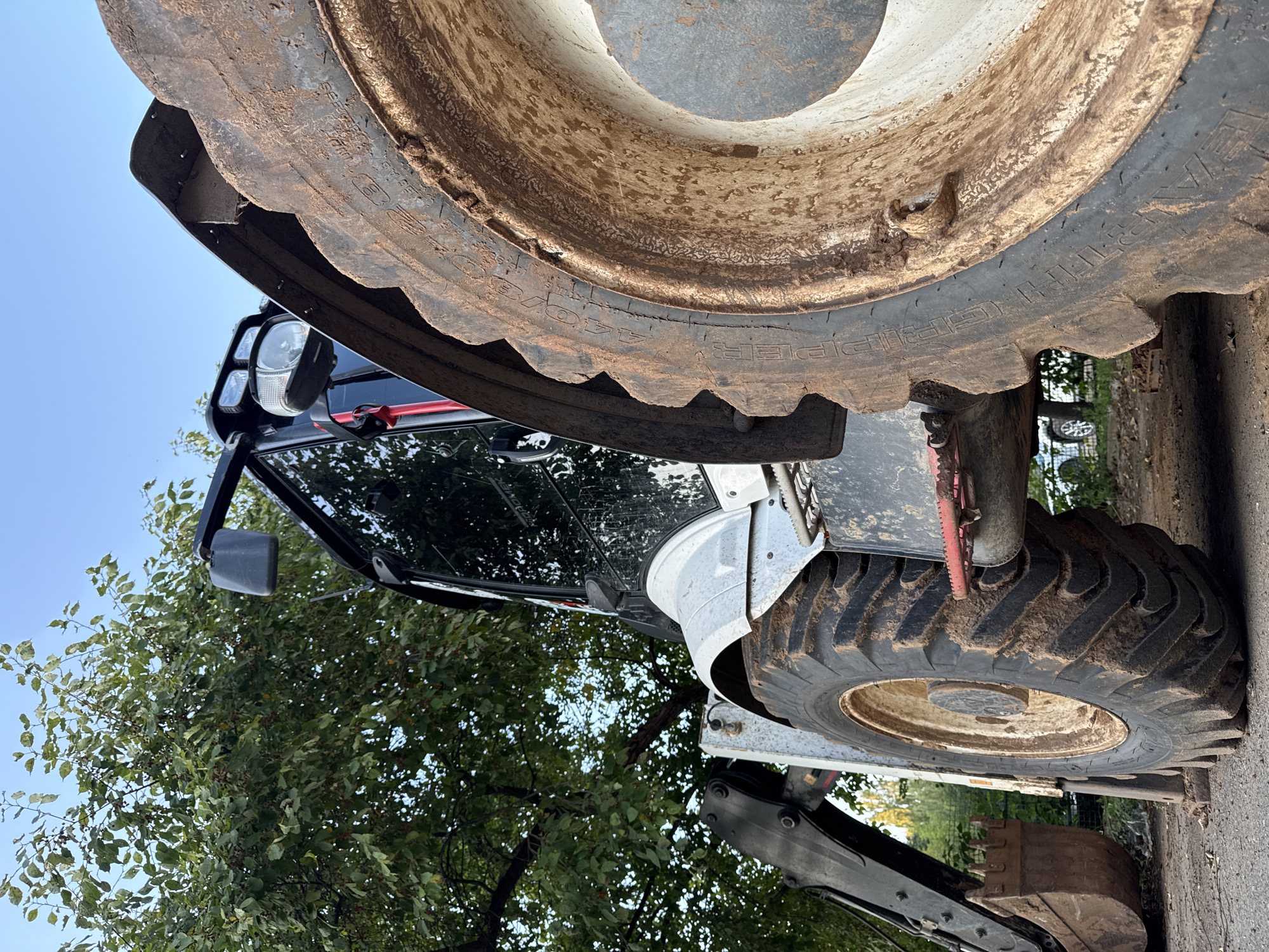This is a low-angle, close-up shot of a dirty backhoe loader parked outdoors on a paved surface, possibly a driveway or lot. The vehicle is predominantly white and black, with large, mud-caked tires. To the left, a portion of the loader's arm and a dirt-filled bucket are visible. Behind the vehicle, lush green trees with some red berries are present, with a clear blue sky above. The scene appears to be from Izhevsk, Russia, as indicated by the metadata. There are no people or animals present in the image.