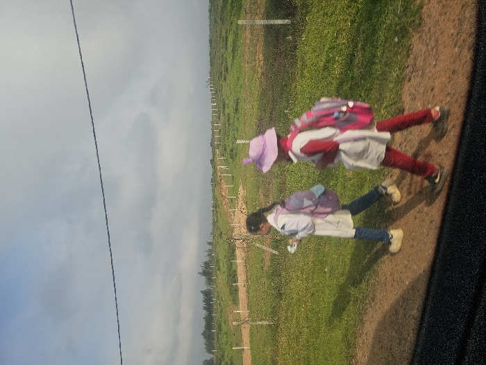 Two young females, appearing to be students, are walking right-to-left along a dirt path in a rural area of Ameur عامر, Morocco. The subject on the left wears a white smock over blue jeans and light-colored shoes, carrying a purple backpack and holding a small white object in her right hand. The subject on the right, slightly behind, wears a white smock over red trousers and dark shoes, a light purple wide-brimmed hat with two protrusions, and a pink and purple backpack. The environment consists of a green grassy field on either side of the path, marked by a line of white fence posts extending into the distance. A treeline is visible on the far-left horizon. The sky is overcast with a mix of light blue and grey clouds, and a dark utility line stretches horizontally across the upper frame. Shadows indicate daylight from the left.