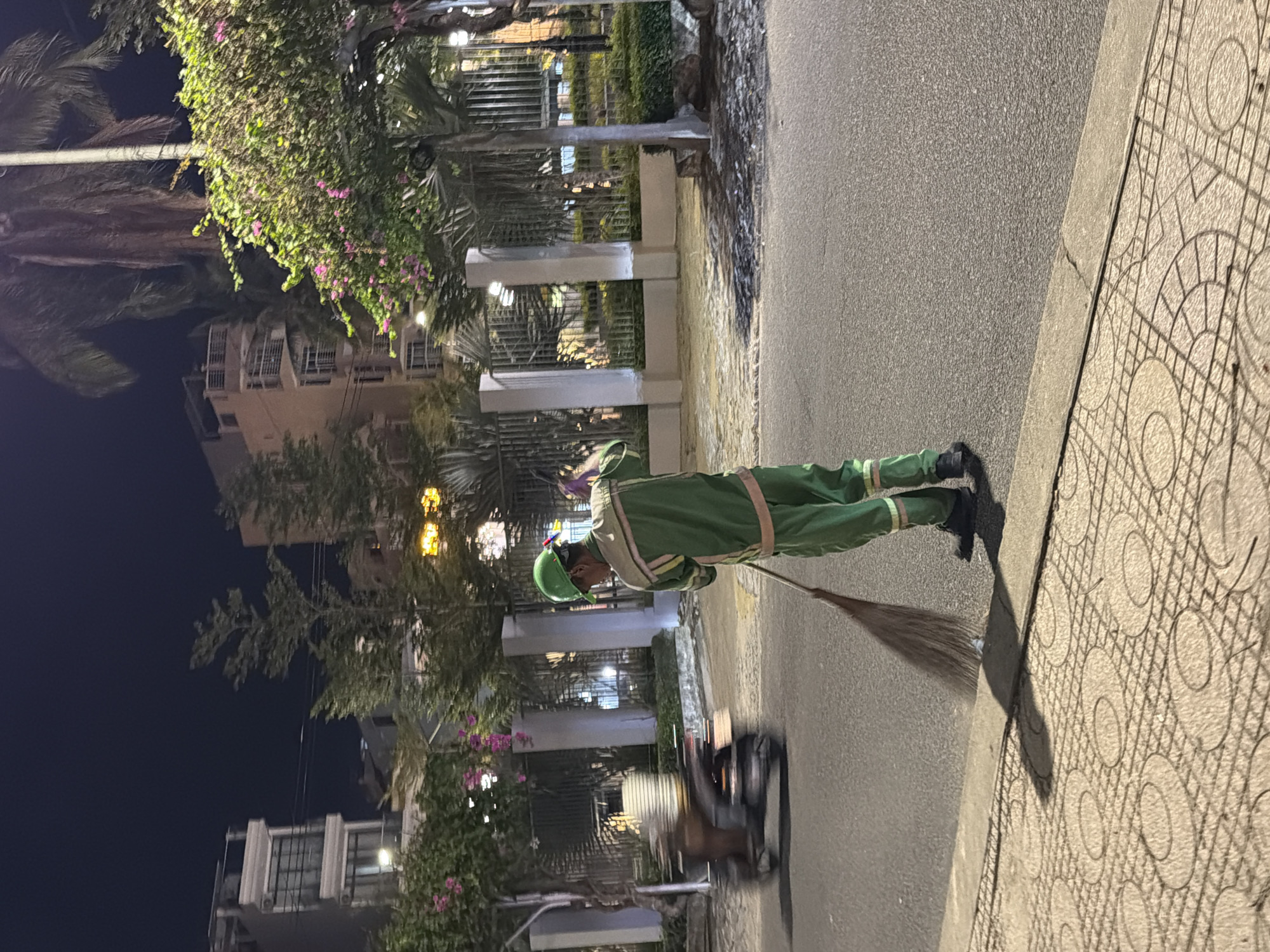 A street cleaner in a green uniform and helmet sweeps the pavement at night in Nha Trang, Vietnam. The street cleaner is on the right side of the image, facing away from the camera and toward the left. They are using a broom to sweep debris from the sidewalk onto the street. In the background, there are buildings, trees, and streetlights. A motorcycle is blurred in motion on the left side of the image. The overall scene depicts a typical urban environment at night, with a focus on the street cleaning activity.