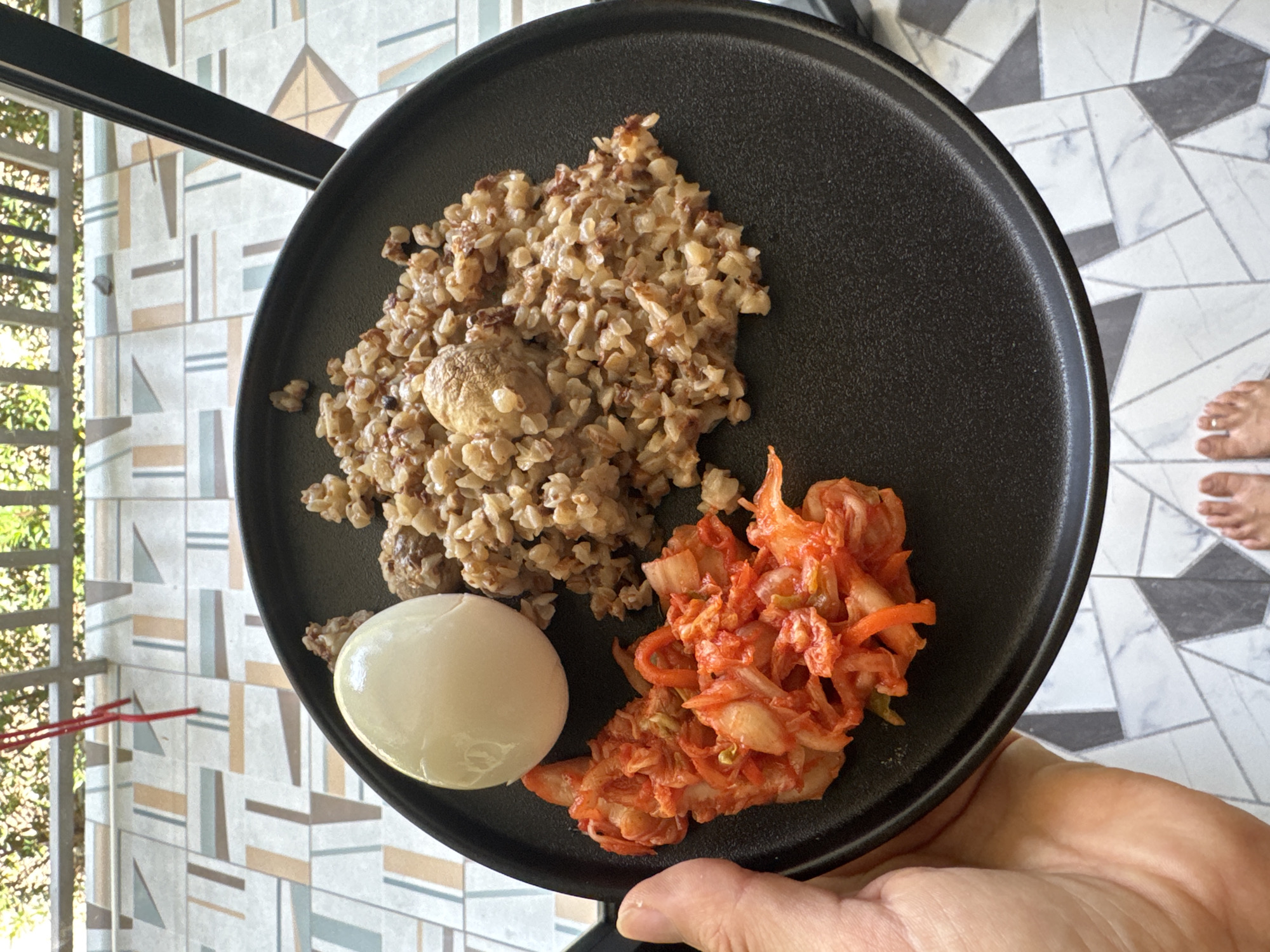 A black plate is being held in a person's hand. On the plate is a pile of cooked buckwheat, a boiled egg, and kimchi. The floor has patterned tiles. A glimpse of a balcony with plants and a lattice-like structure is seen. The location is Moo 4, Thailand. The image suggests a meal being held or presented. The person's toes are visible in the upper right.