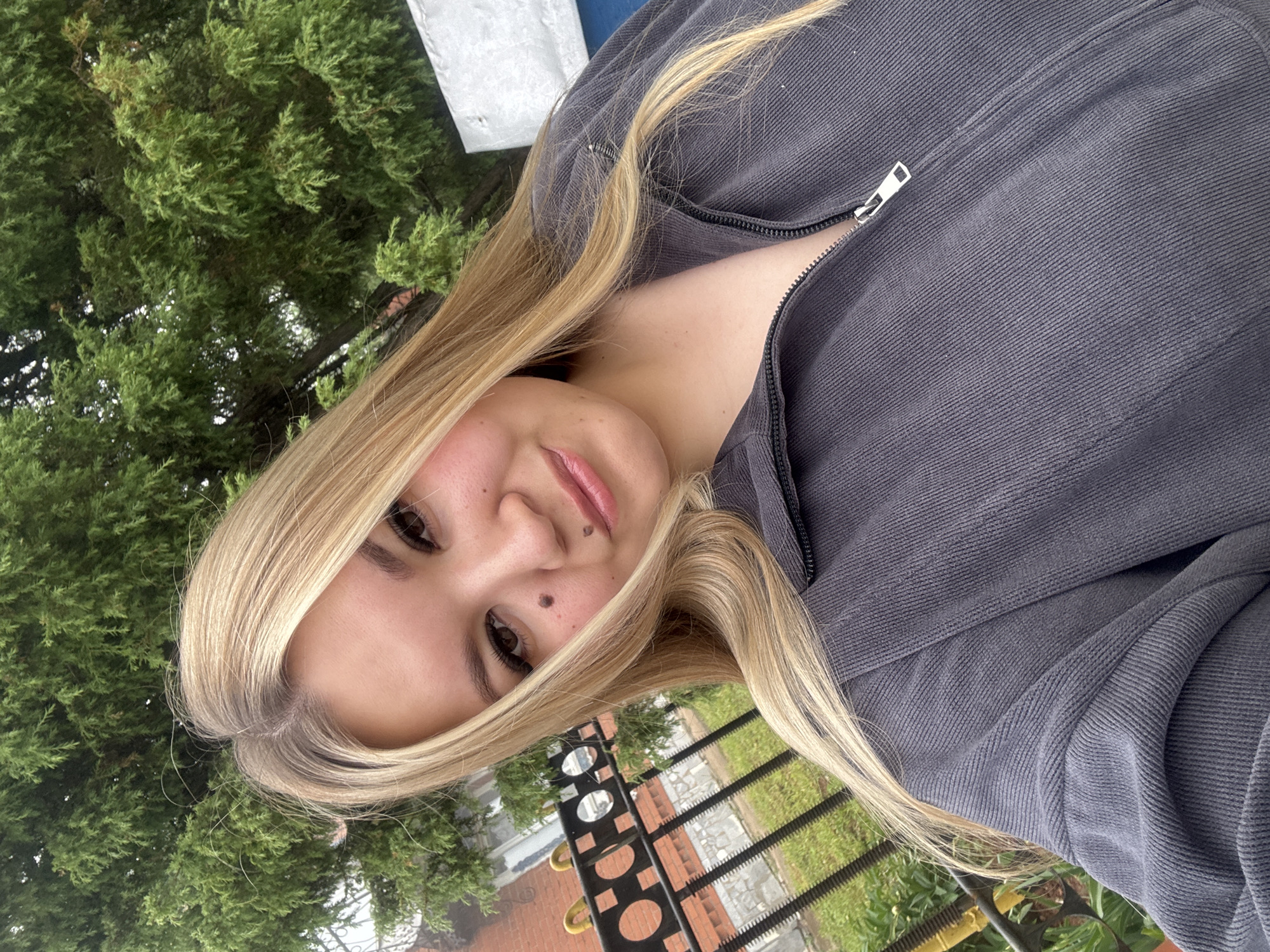 A close-up, highly tilted portrait captures a young woman outdoors in Большой Букор, Russia. Her head and torso are positioned diagonally, with her long, straight blonde hair, parted on the side, cascading over her shoulders. She looks directly at the camera with a soft smile, revealing dark eyes, well-defined eyebrows, and beauty marks on her right cheek and above her upper lip. She wears a dark gray, possibly corduroy or wide-rib knit, zip-up jacket, partially unzipped. The background features lush green foliage from a dense tree or bush filling the upper left side, contrasting with a black metal fence or railing visible in the lower right, beyond which hint of grass and possibly a building are discernible. The natural lighting indicates a daytime setting. There is no visible text in the image.