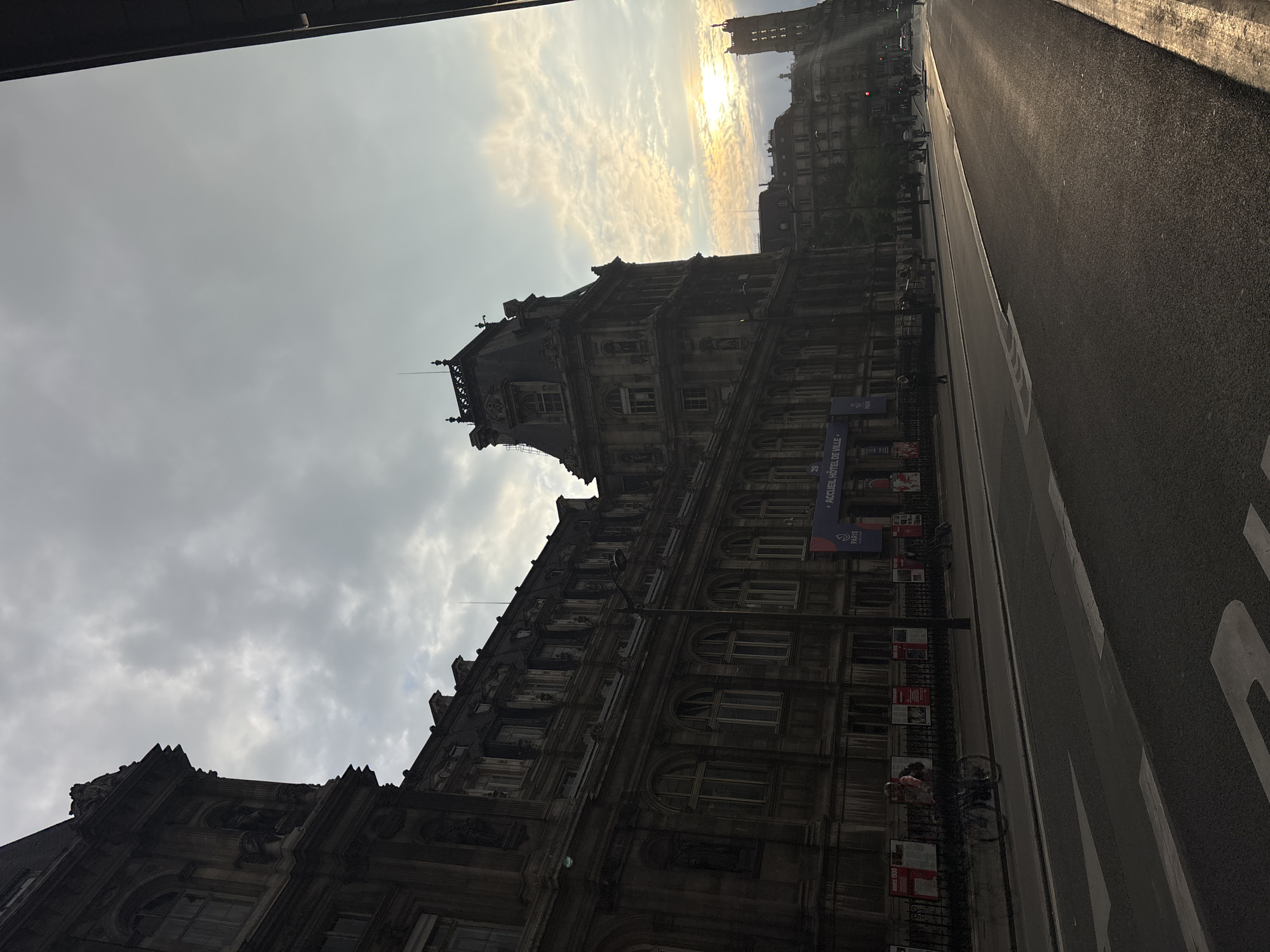 The image shows the Hôtel de Ville (City Hall) in Paris, France. It is an overcast day, but the sun is trying to break through the clouds. The building is a large, ornate structure with many windows and a tower. There is a street running alongside the building, with a pedestrian walkway between the building and the street. Bicycles are parked along the walkway. There are a few people in the scene. Banners and posters are attached to the railings next to the road, advertising an event. The camera is positioned at a tilted angle, adding to the perspective of the towering building.