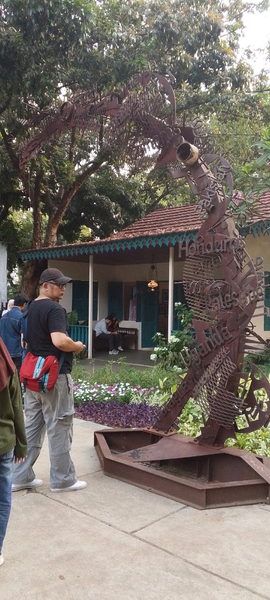 This image captures an outdoor scene near a traditional Indonesian house in Central Jakarta. The primary focus is a large, metallic sculpture of a seahorse, intricately crafted from what appears to be rusted metal. The sculpture is positioned on a concrete surface, adjacent to a garden with various flowers and plants.
In the foreground, a man wearing a black t-shirt, grey cargo pants, and a black baseball cap is standing and looking to his left. He has a red and blue satchel slung across his chest. Behind him, partially visible, is a person wearing a green jacket and blue jeans.
Further back, in front of the house, are several individuals. Two people are seated on a bench on the porch of the house, one of whom is using a mobile phone. The house itself features a tiled roof with decorative trim and green shutters. The environment suggests a public space, possibly a museum or historical site, given the presence of the sculpture and the traditional architecture. Trees with lush green foliage provide a natural backdrop to the scene. The overall atmosphere is relaxed and casual, indicating a daytime setting.