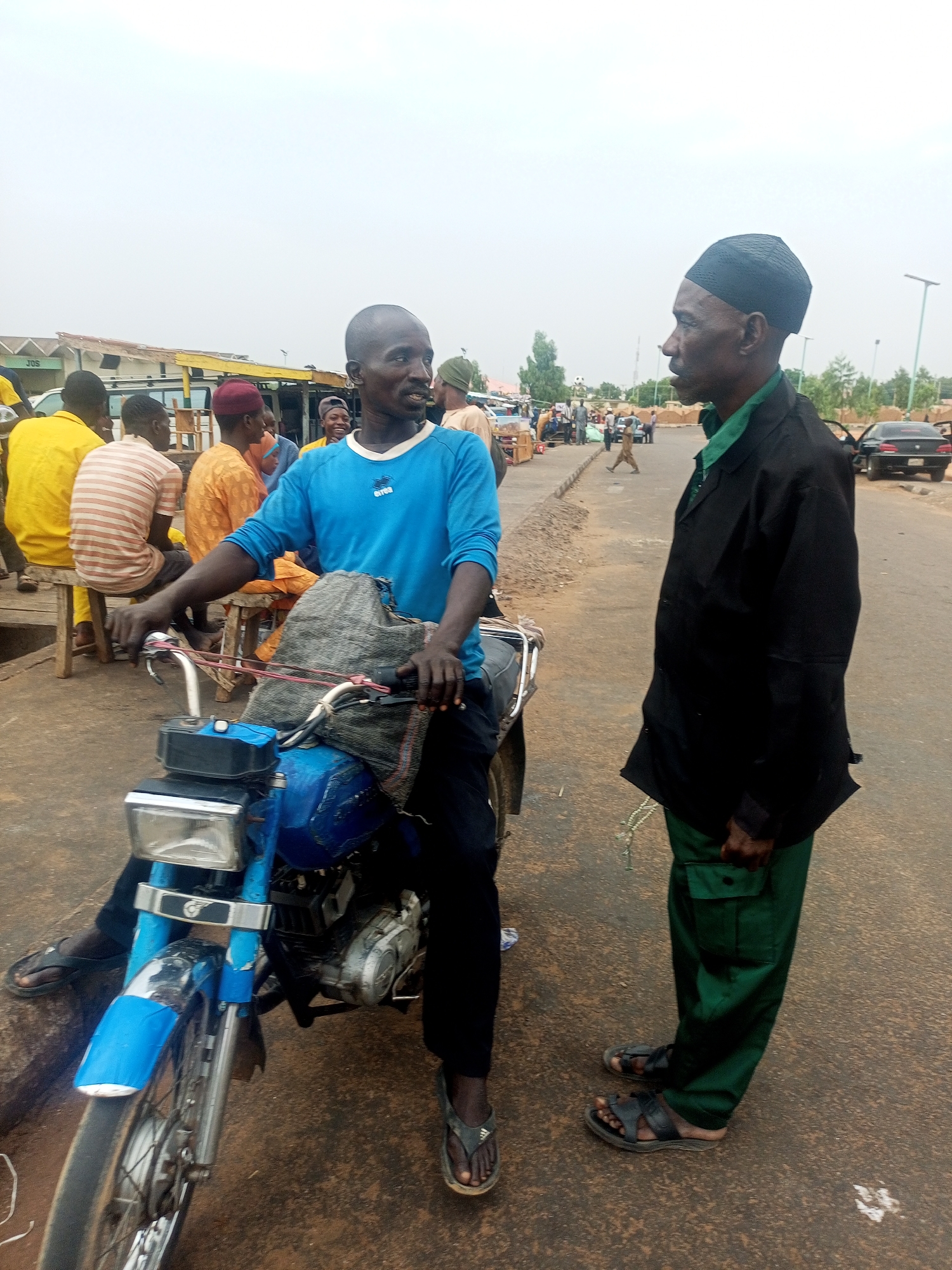 The image captures a scene in Gombe, Nigeria. A man in a blue shirt is seated on a blue motorcycle. Another man, wearing a black cap, green shirt, black jacket, and green trousers, stands facing him. Both men are wearing sandals. The motorcycle is positioned on a paved road or street. Behind them are other people, stalls, and buildings, indicating a market area. A car is parked nearby. The sky is overcast.