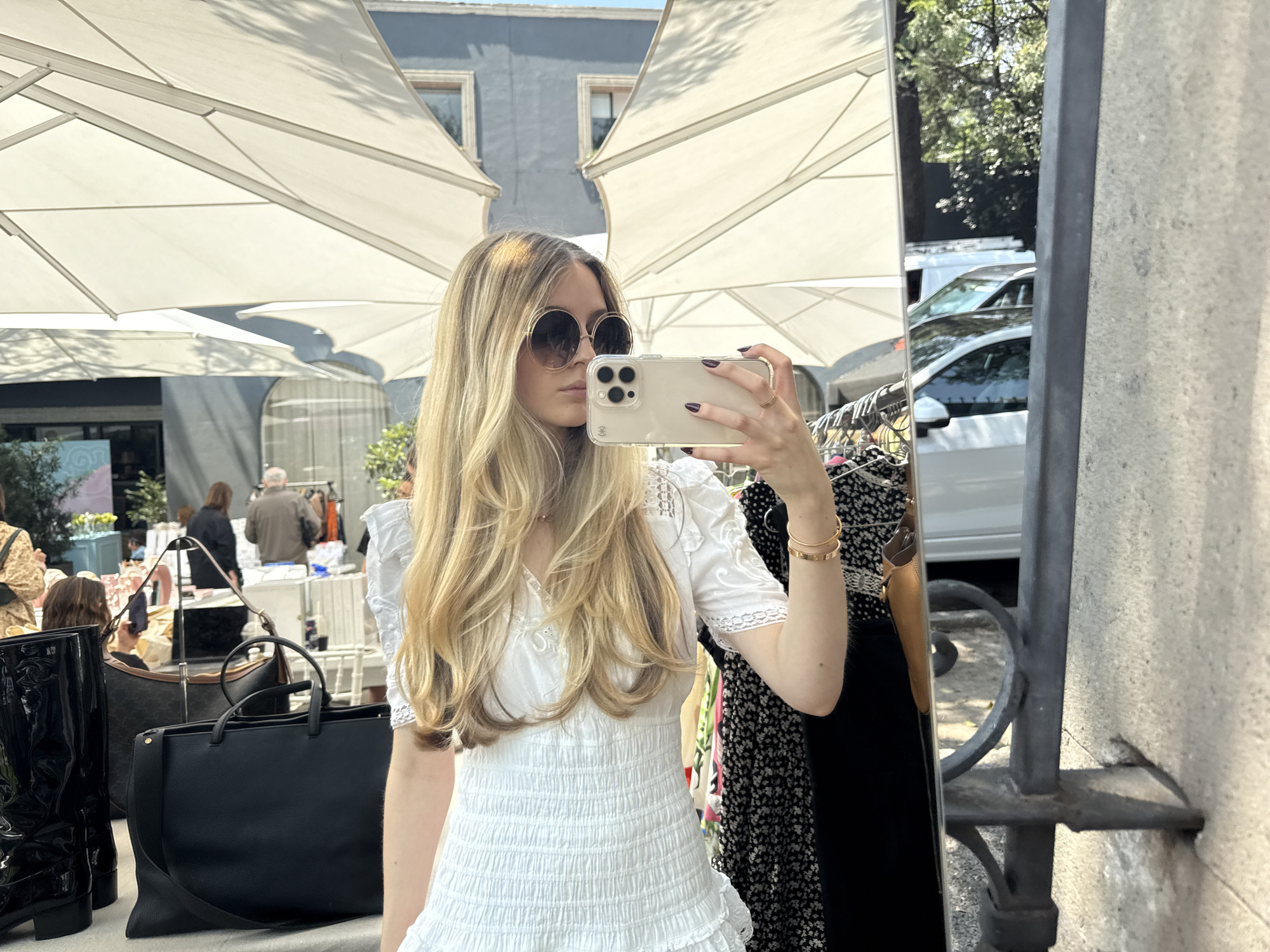 A woman stands in front of a mirror, taking a selfie with her smartphone. She is wearing large sunglasses, a white dress, and gold bracelets. Her long blond hair is styled in loose waves. In the background, there are white market umbrellas and blurred figures of other people, suggesting an outdoor market setting. There are also handbags and boots visible in the foreground, indicating a retail context, likely in Mexico City, Mexico. Cars and a building can be seen reflected in the mirror.