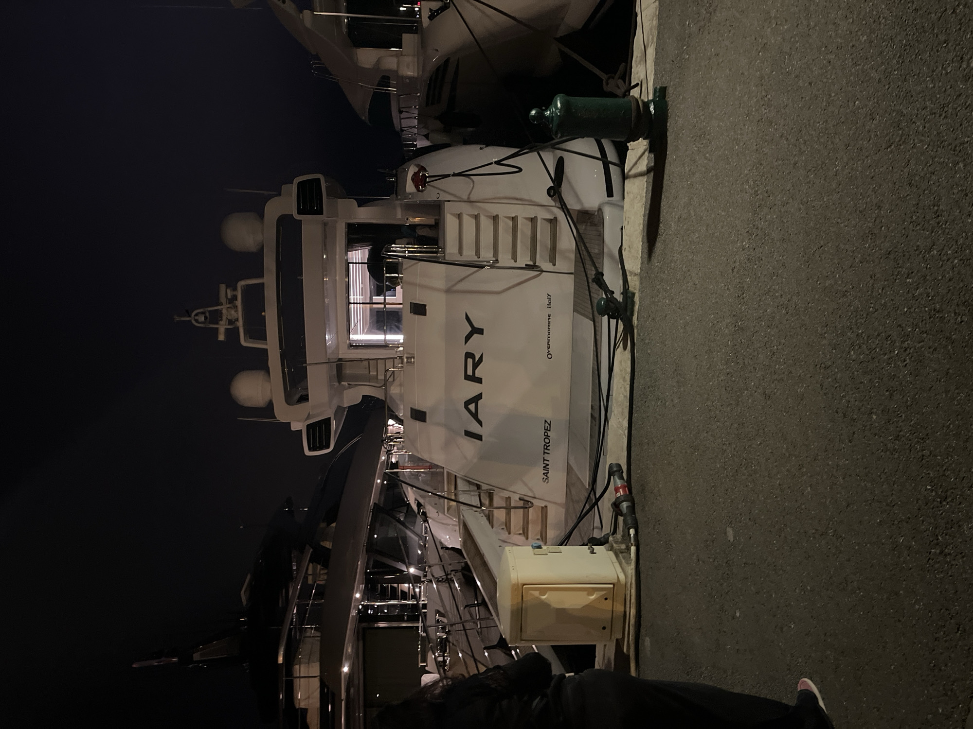 At night in Grimaud, France, a large yacht named "IARY Saint Tropez" is docked alongside a quay. The stern of the yacht features stairs leading up to the deck. The yacht is white with black lettering and is adjacent to a textured concrete quay. A portion of a person is visible in the lower part of the image.