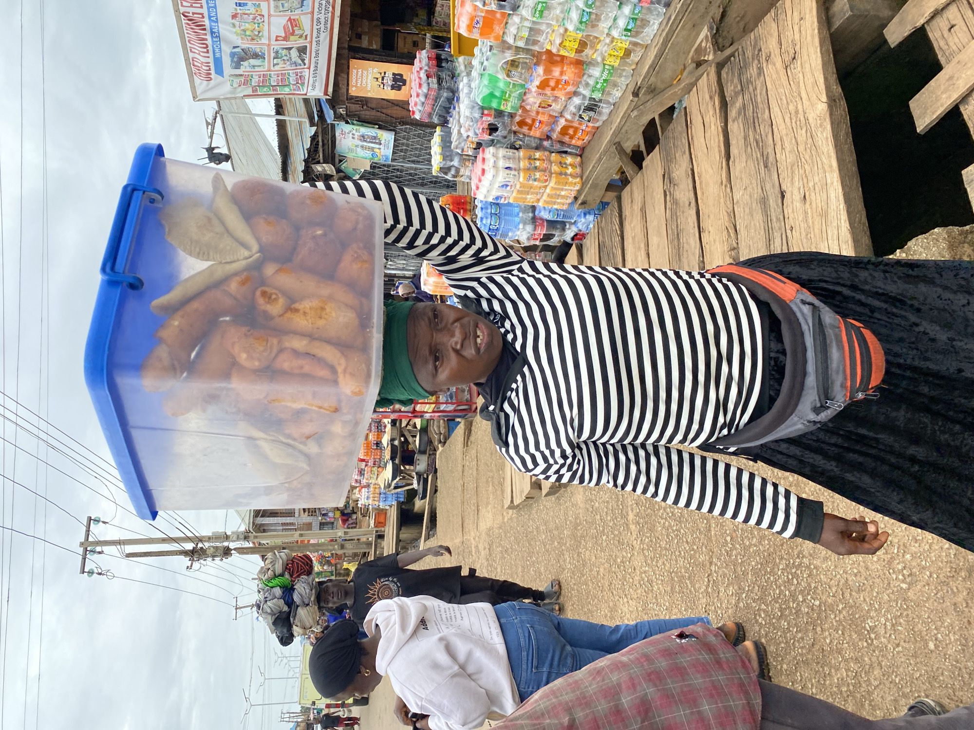 In Bukuru, Nigeria, a woman with dark skin and a green headscarf balances a large, clear plastic tub filled with food items on her head. She wears a black and white striped long-sleeved shirt and a black skirt, with an orange and gray fanny pack around her waist. Her mouth is open as she looks forward, her teeth visible. She stands on a wooden walkway in what appears to be a market. To her right, a wooden stall is laden with numerous bottles of beverages, including brands like Pepsi, Sprite, Fanta, and Coca-Cola, as well as some bottled water and yogurt drinks. In the background, a man wearing a black t-shirt and dark pants walks with a load of items on his head, and other people are visible in the distance. Electrical wires and poles are prominent in the sky, suggesting an urban or semi-urban environment. A sign in English is visible above the beverage stall. The ground is a mixture of gravel and sand.