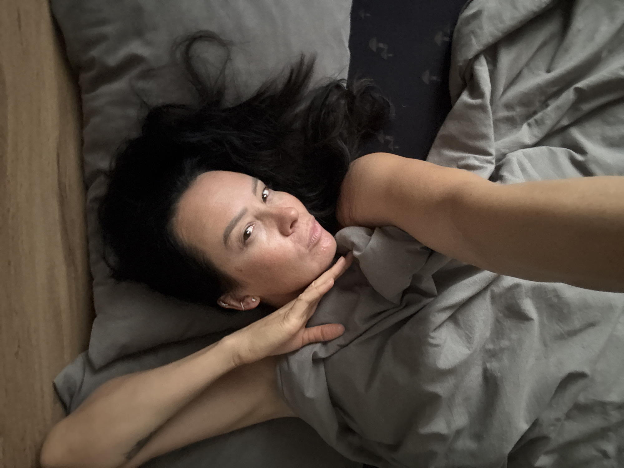 A woman lies on a bed covered with gray linens. Her dark hair is spread across the pillow. She is partially covered by a gray blanket, holding her hand to her chin. The background includes a wooden surface, likely a headboard or bedside table, and a dark-patterned pillow or part of the bedding. The location, Chaikovsky, Russia, is extraneous and cannot be verified from the image.