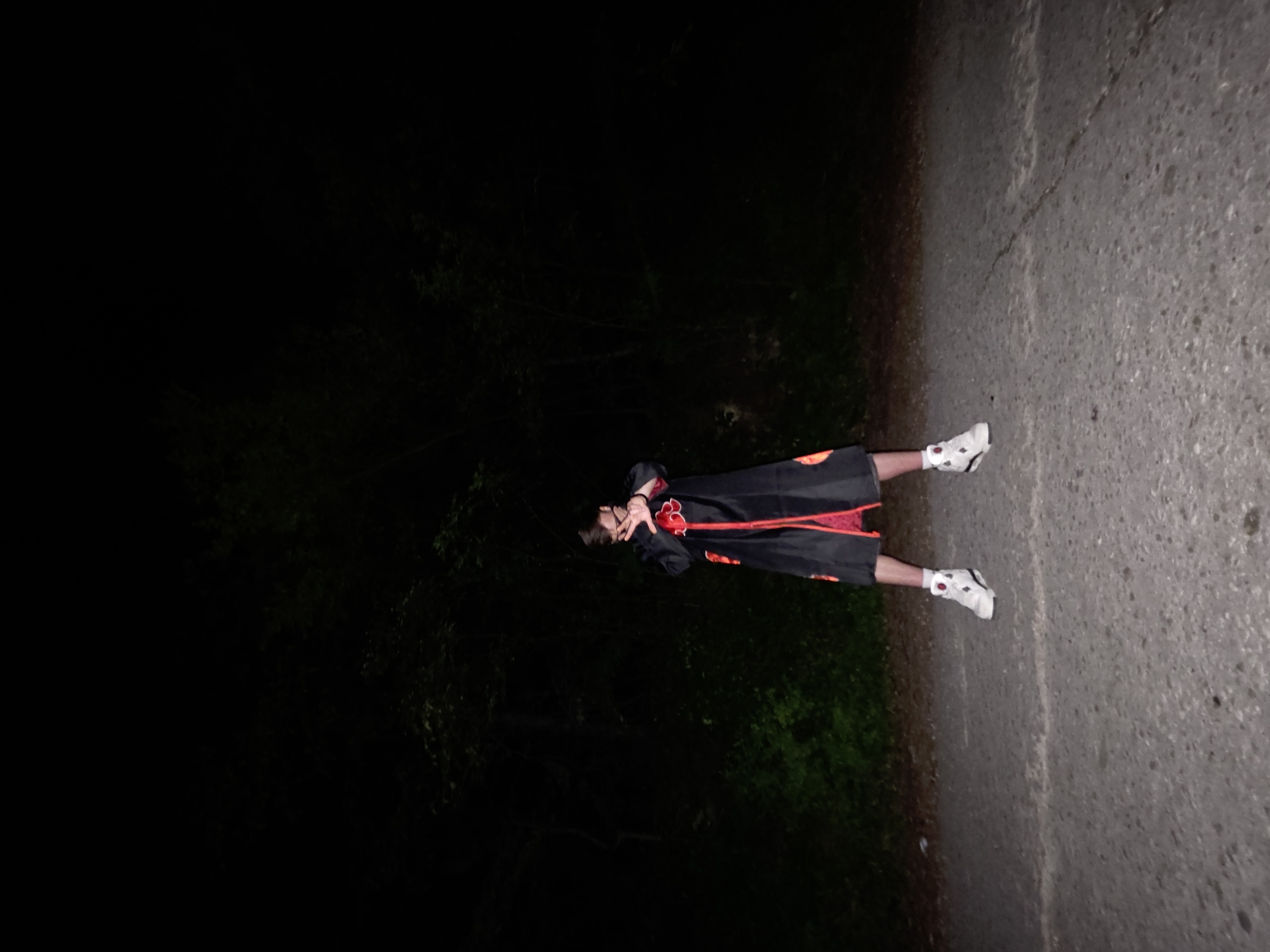 A young person is positioned on the right side of a concrete pathway near a wooded area. They are dressed in an Akatsuki cloak and white sneakers. The left side of the image shows a dark, wooded area. The location is Lyaskovo, Bulgaria.