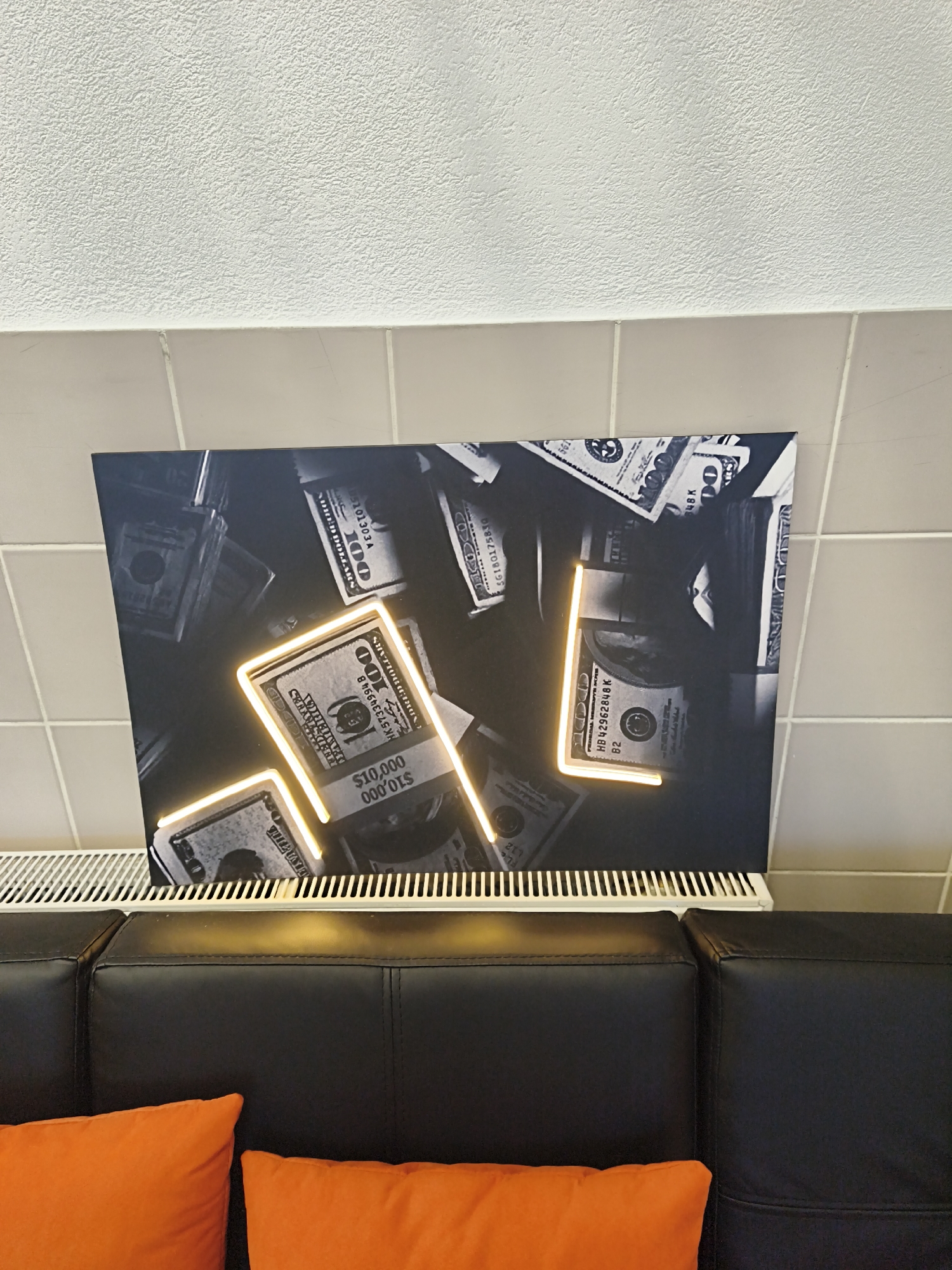 A framed black and white photographic artwork of US dollar bills, some stacked and some as individual notes, is displayed on a wall above a white radiator and black leather couch. The artwork features glowing neon orange outlines of stylized shapes that partially obscure some of the money. Two bright orange throw pillows rest on the couch in the foreground. The wall above the artwork is textured and white, while the wall beneath it consists of light beige square tiles. The image was taken indoors, likely in a residential or commercial space. There are no people or discernible actions taking place. The scene is static and presented from a slightly elevated perspective. The artwork is the primary subject of the image, with the furniture and decor serving as context. The image does not provide any information about the city of Minsk, Belarus.
