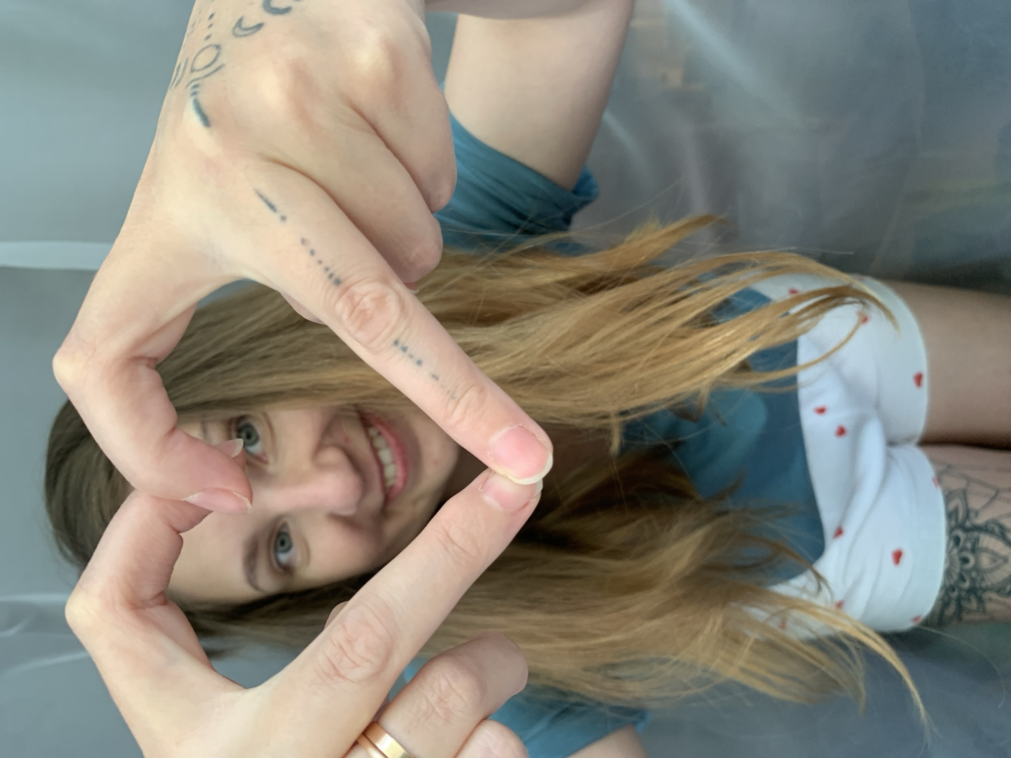A woman is lying on her back, smiling and looking through a finger frame she is making with her hands. She has blonde hair and blue eyes. She is wearing a blue t-shirt and white shorts with red hearts. Her left hand has tattoos, including crescent moon shapes and a line of dots. She has a gold ring on her left ring finger. A tattoo is visible on her right thigh. The location is unknown and there is no evidence provided to suggest it is Chaikovsky, Russia. The background appears to be a smooth white or grey surface.